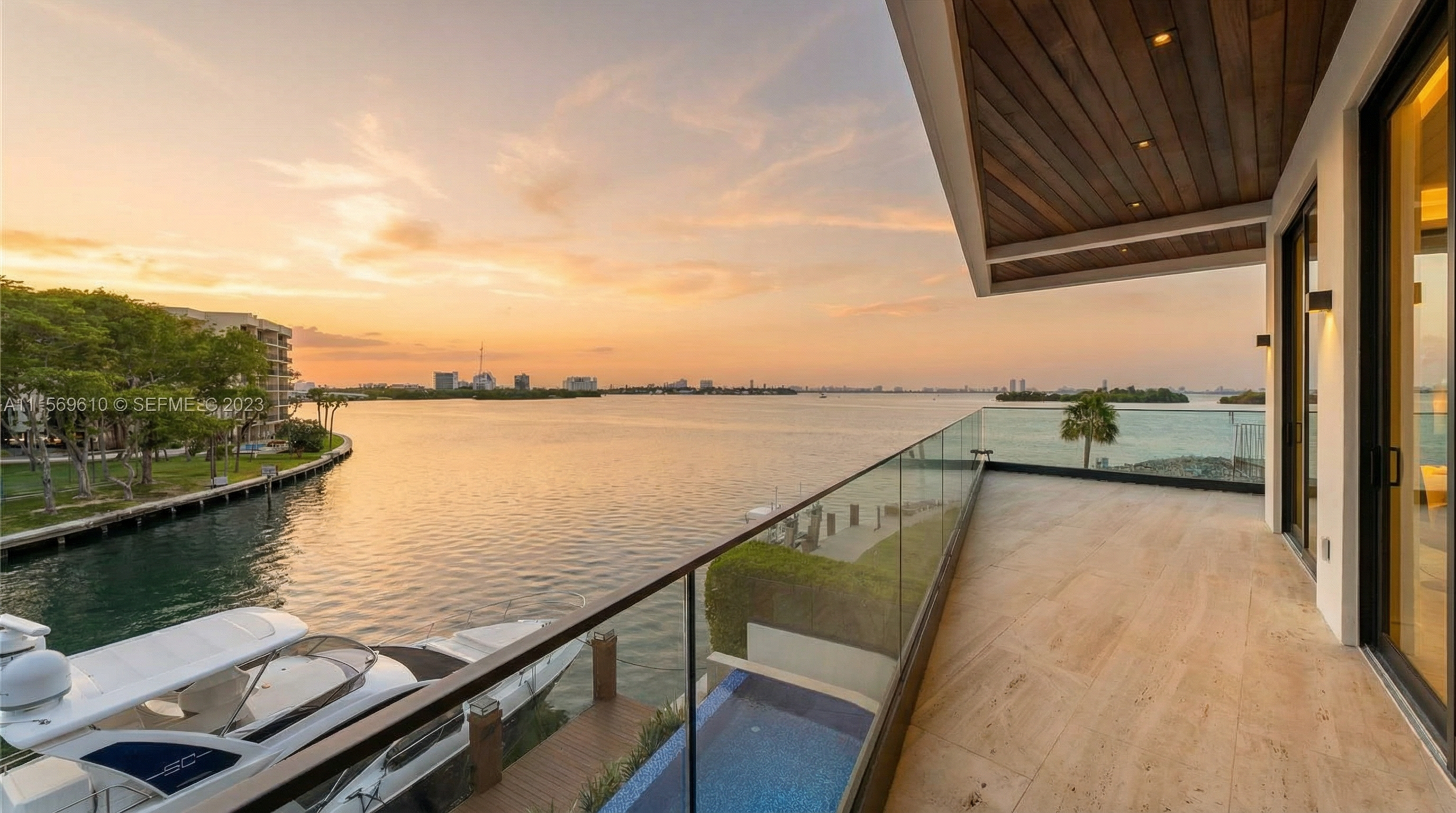 This modern, high-end balcony offers expansive, panoramic views of the waterfront with a sunset sky. The space features stylish travertine-style tiling, a sleek glass railing, and a warm wood-paneled ceiling with recessed lighting. Below, a private boat slip and small pool area add to the luxury appeal of this prime real estate property.