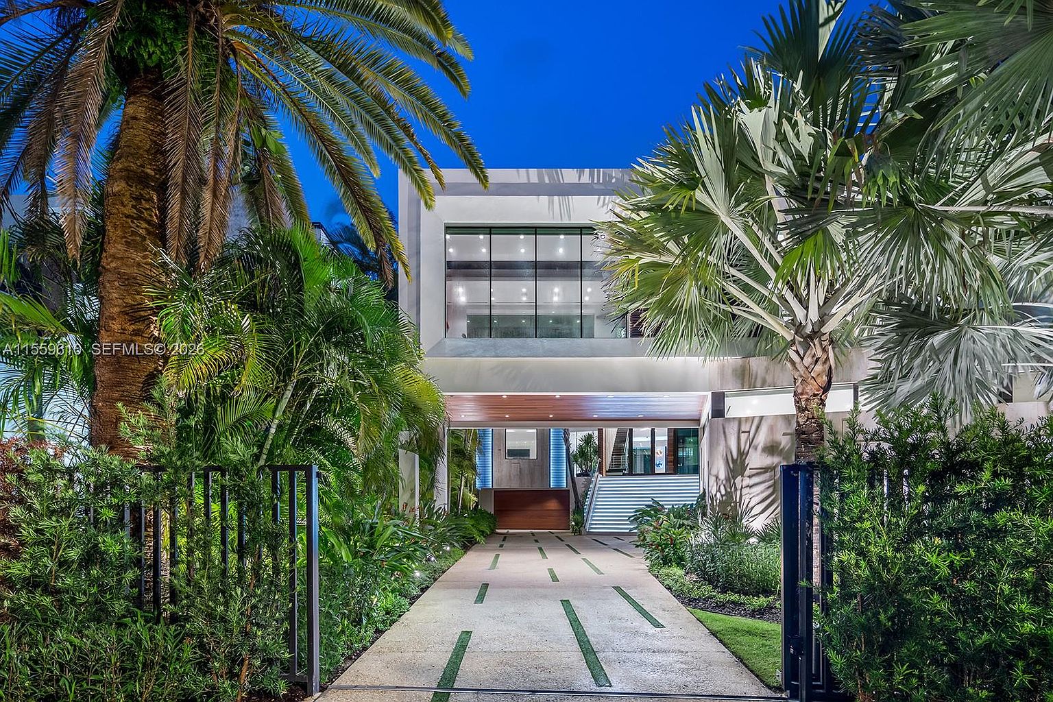 This image showcases the grand entryway of a modern home, framed by lush tropical landscaping and palm trees. A long, paved driveway leads to the entrance, flanked by manicured greenery and a sleek, black metal gate. The home's facade features a large window and a covered entryway, creating a welcoming and luxurious first impression.