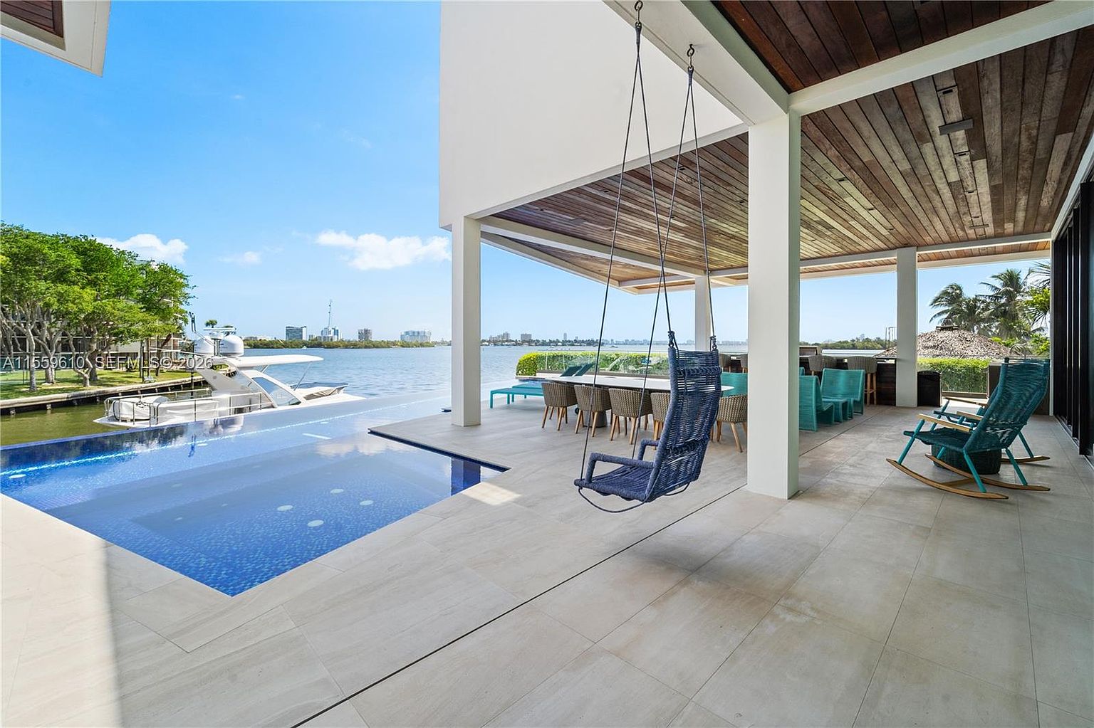 This image showcases a luxurious outdoor patio area with a modern design. The space features a covered area with wooden ceilings, a hanging swing chair, and a dining table with chairs. An infinity pool extends towards the waterfront, offering stunning views of the water and cityscape in the distance, creating a seamless blend of indoor and outdoor living.