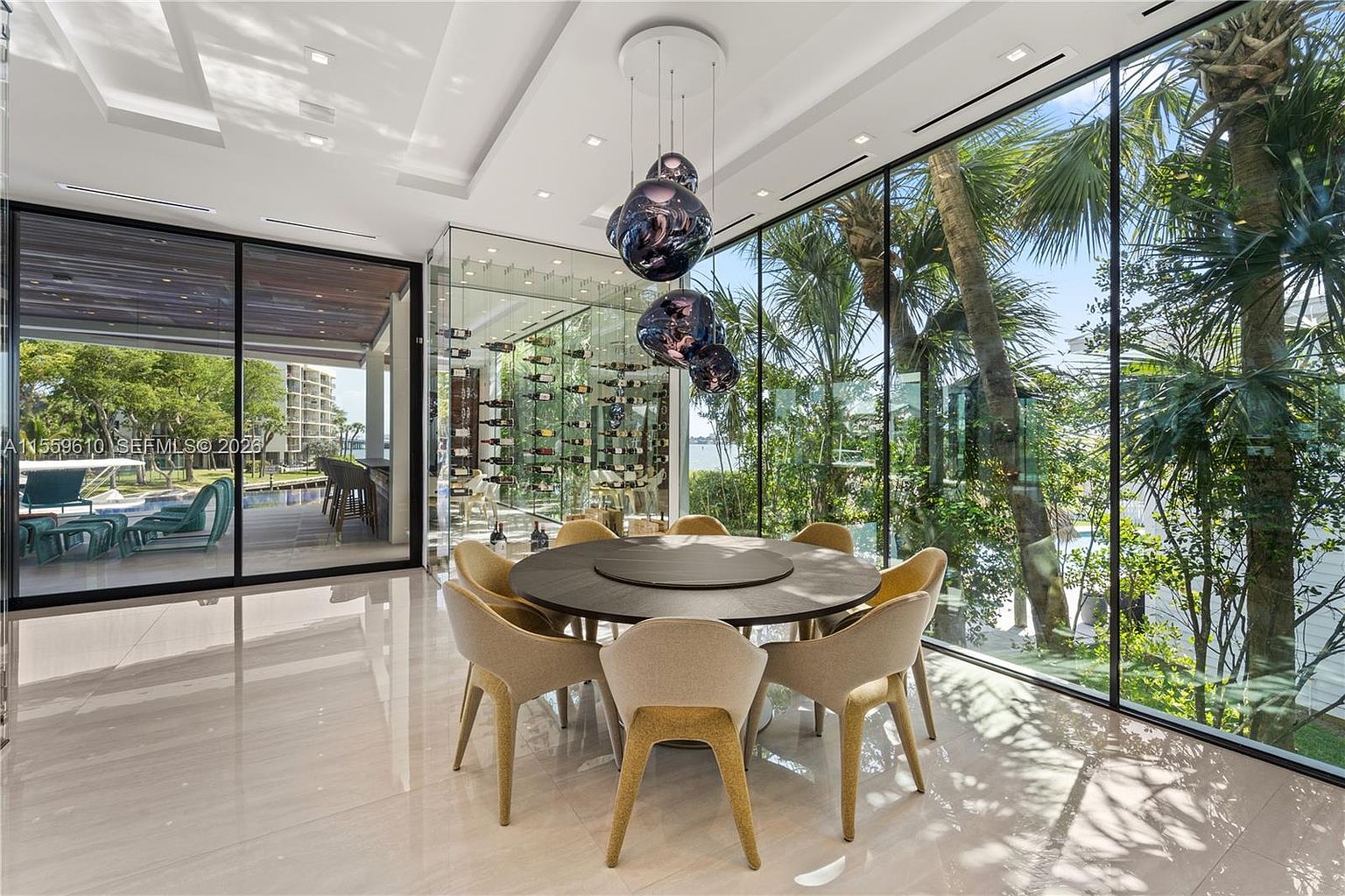 This is a modern dining room featuring a round dining table with eight chairs, a glass-enclosed wine cellar, and floor-to-ceiling windows offering views of lush greenery. The room is illuminated by a unique pendant light fixture, and the polished tile flooring reflects natural light, creating a bright and airy atmosphere. The perspective is from the corner of the room, capturing the dining area and the view outside.