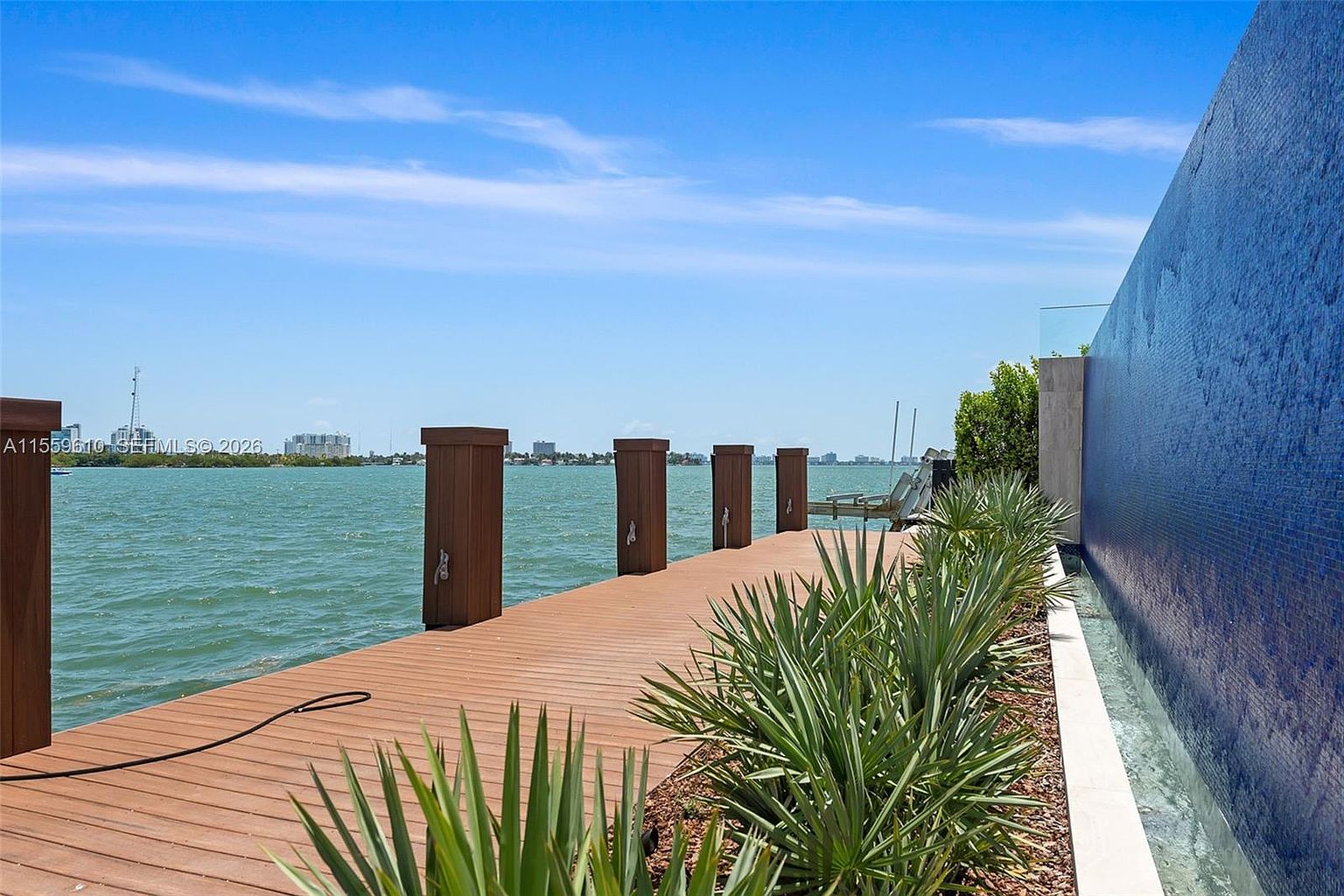 This image showcases a waterfront deck featuring wooden planks and sturdy wooden posts along the edge. Lush green plants line the side of the deck, adding a touch of nature. A unique blue tiled wall with a water feature provides a modern and luxurious backdrop, enhancing the property's appeal.