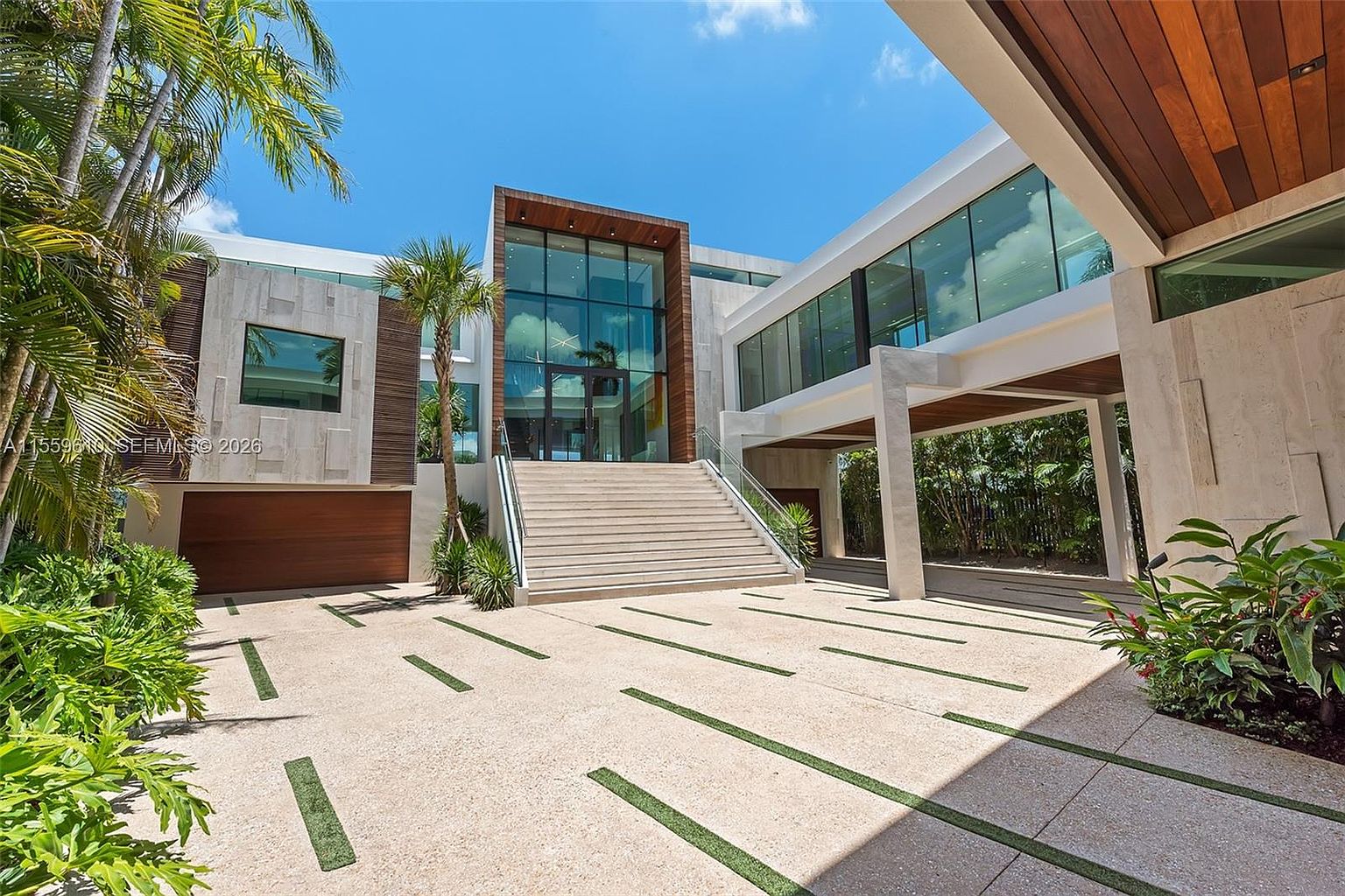 This is a striking front view of a modern luxury home. The architecture features a grand entrance with a large glass facade, complemented by stone and wood accents. A wide staircase leads to the entrance, and the driveway is designed with integrated grass strips, adding a touch of greenery to the property's curb appeal.