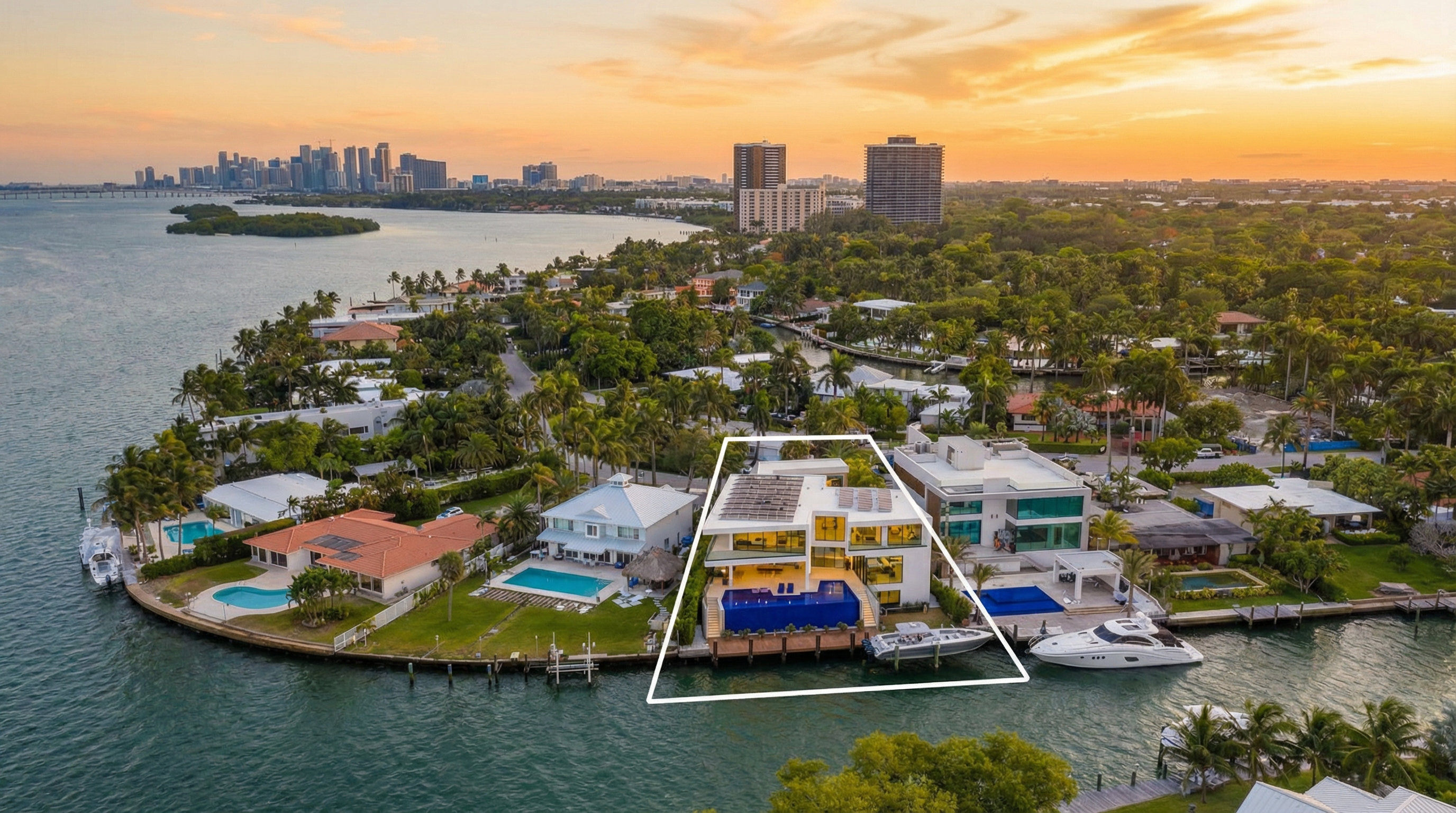 This stunning aerial view highlights a modern, luxury waterfront estate in a coastal setting at sunset. The property features a prominent multi-level design, a rooftop solar panel array, and a striking glass-fronted infinity pool overlooking the bay, accompanied by a private boat dock. Its prime location on a point of land offers expansive water views and creates an impression of high-end living in a prestigious neighborhood.