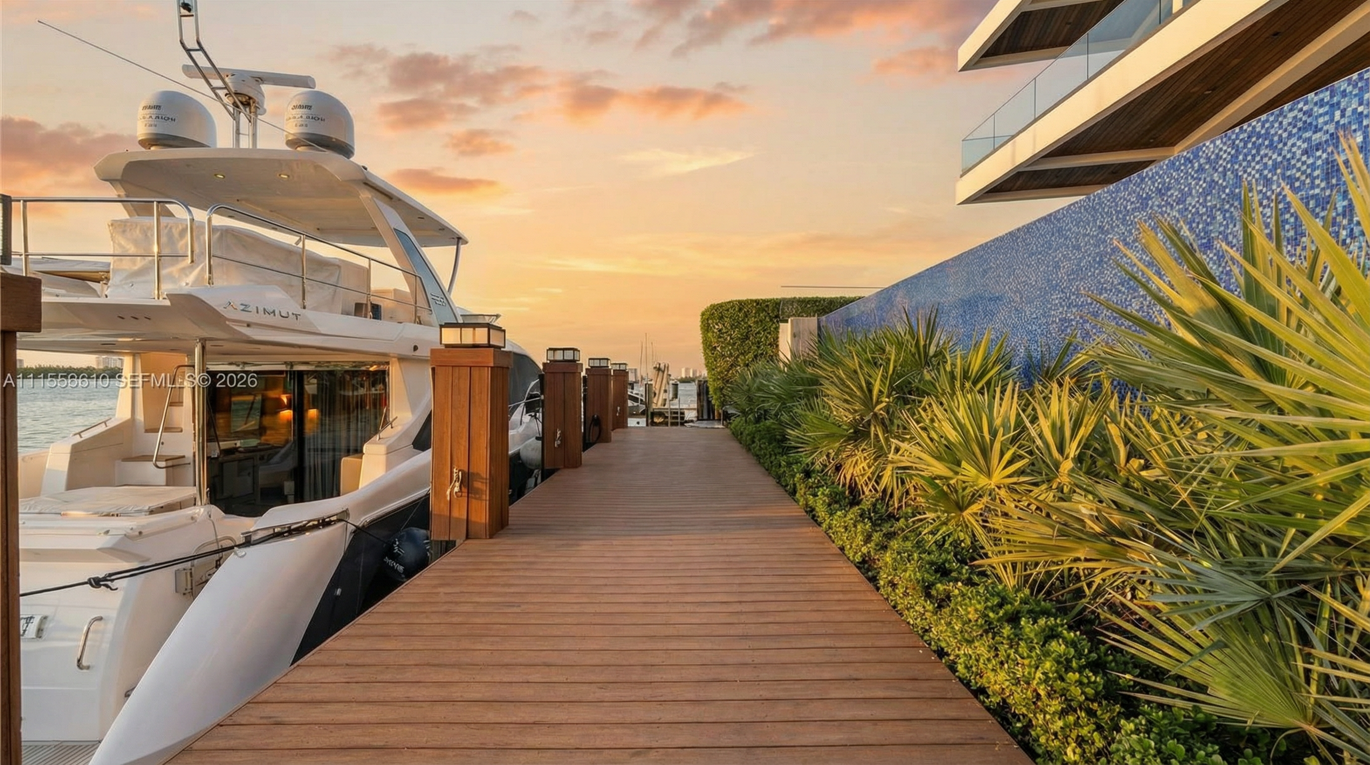 This image showcases a luxurious wooden dock alongside a sleek, docked Azimut yacht during a golden sunset. The waterfront deck features landscaped greenery and a striking mosaic-tiled blue wall, transitioning into modern architectural elements of the property. This setting combines high-end nautical living with sophisticated outdoor design.