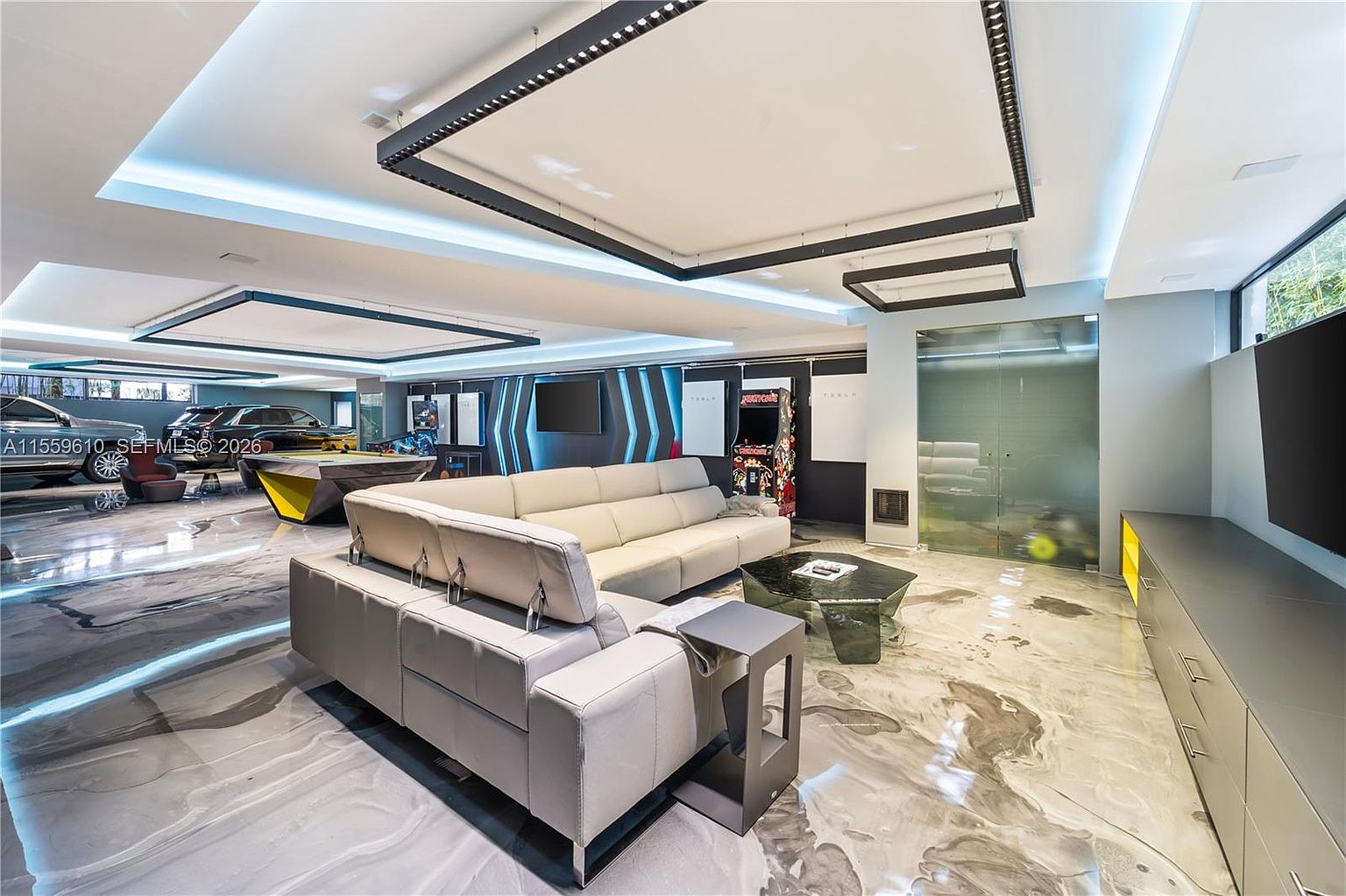 This is a modern and luxurious living room that also functions as a garage. The room features a large sectional sofa, a unique coffee table, and a pool table. The flooring is a striking marble-like material, and the ceiling has recessed lighting and decorative light fixtures. There are also cars parked in the background, adding to the unique design of the space.