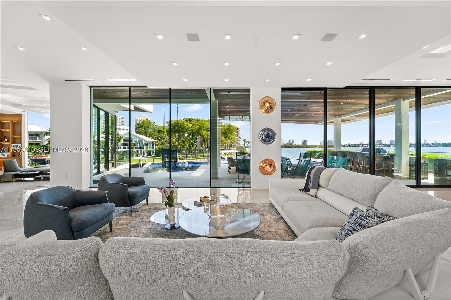 This is a spacious and luxurious living room with floor-to-ceiling windows offering stunning views of the waterfront and outdoor amenities. The room features modern furniture, including a large sectional sofa, armchairs, and stylish coffee tables. The decor is sophisticated and minimalist, creating an open and inviting atmosphere perfect for relaxation and entertaining.