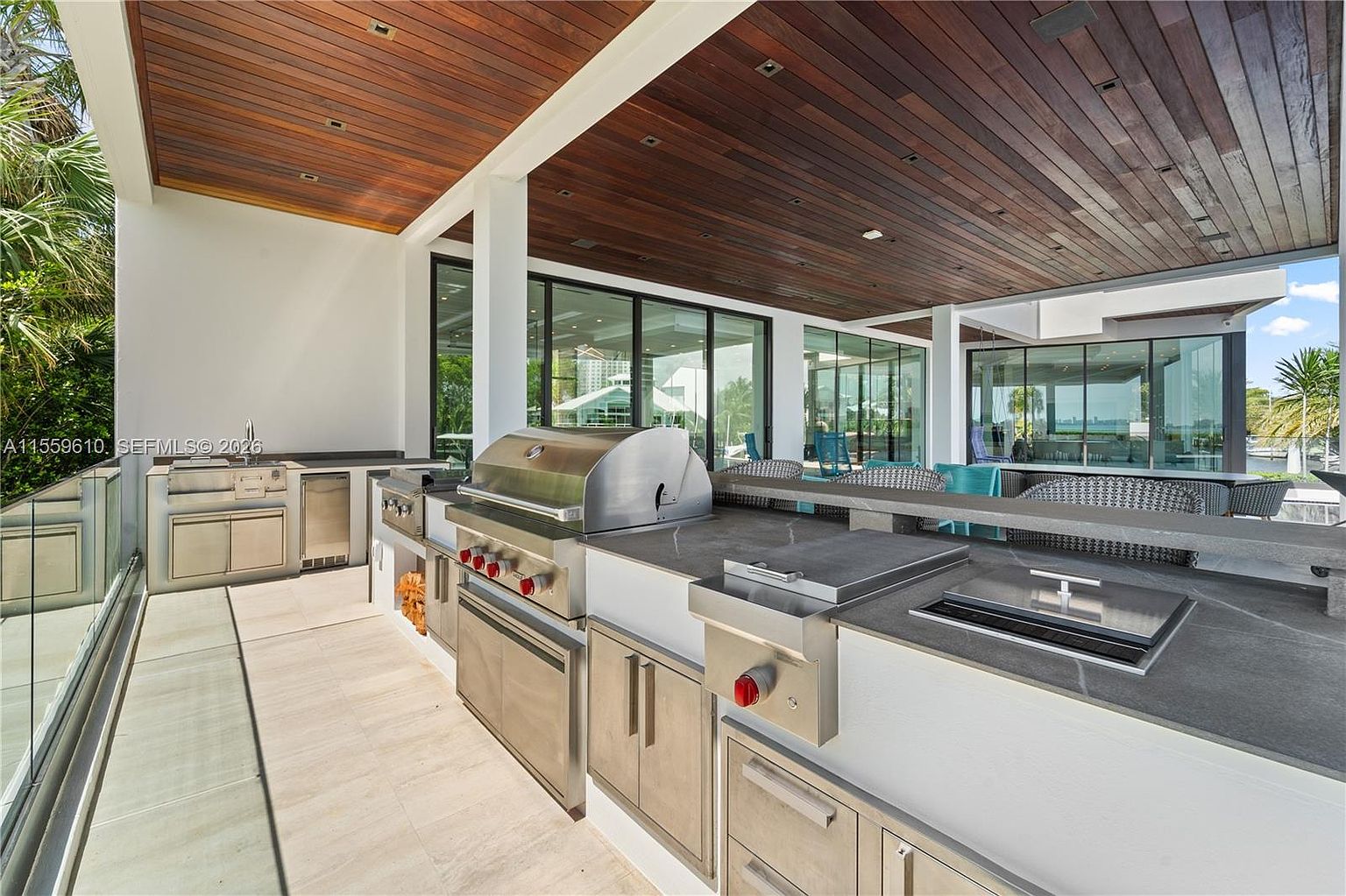 This image showcases an outdoor kitchen area on a patio or deck. The space features stainless steel appliances, including a grill and side burners, built into a modern countertop. The ceiling is finished with wood planks, and large glass doors provide views of the surrounding landscape, creating an inviting and luxurious outdoor living space.