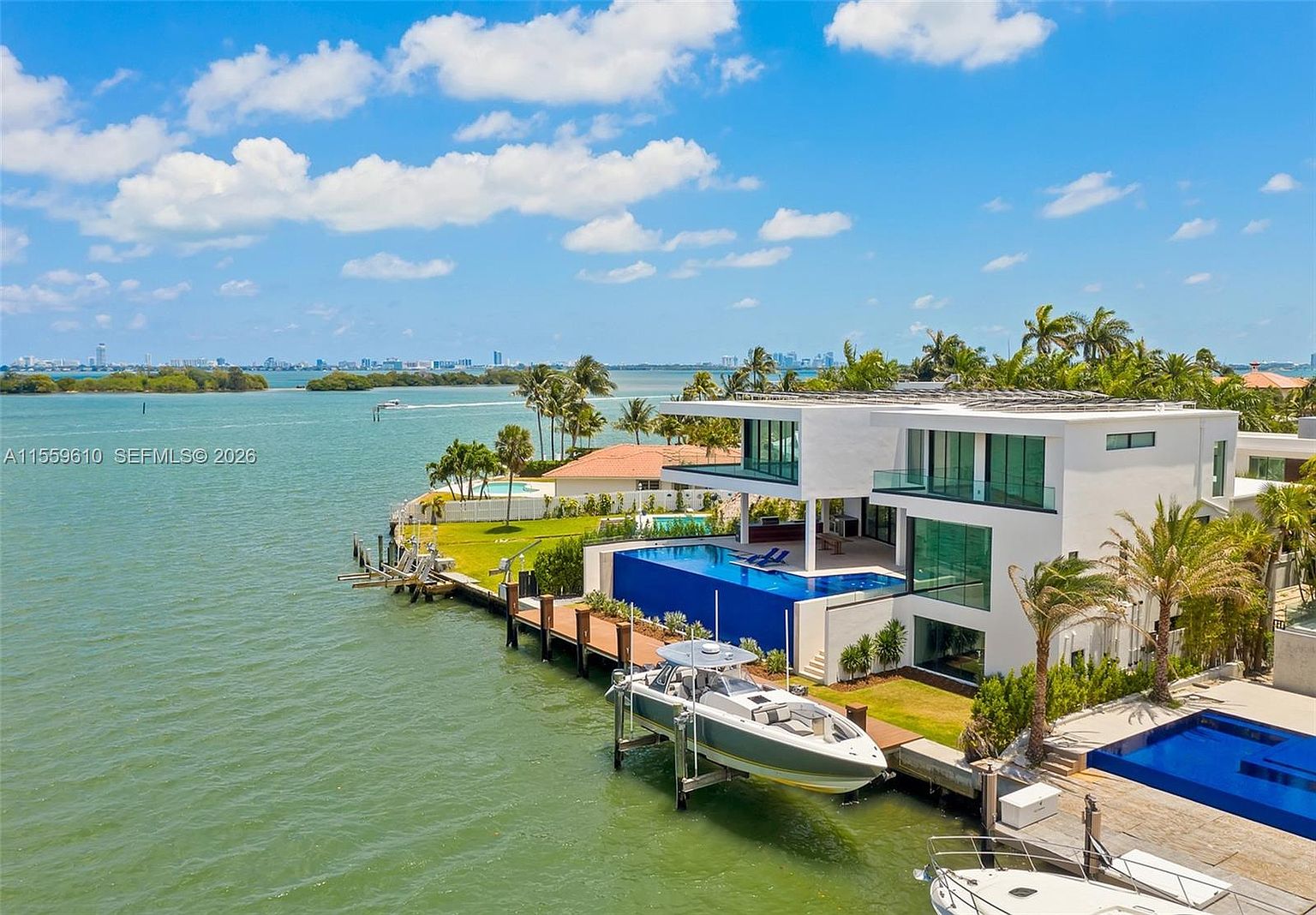 This aerial view showcases a luxurious waterfront property featuring a modern two-story house with a flat roof and expansive glass windows. The property includes multiple pools, a private dock with a boat lift, and lush landscaping with palm trees. The overall impression is one of high-end coastal living with stunning water views.