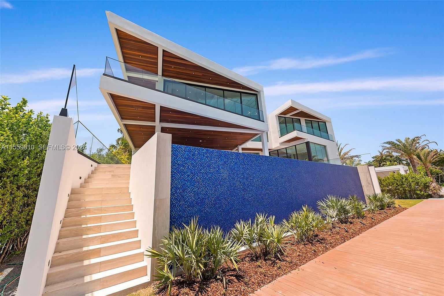 This is a striking front exterior view of a modern home, showcasing its unique architectural design with sharp angles and a combination of white and wood finishes. A vibrant blue tiled wall adds a pop of color, complemented by lush landscaping. A staircase leads up to the house, creating an inviting entryway.