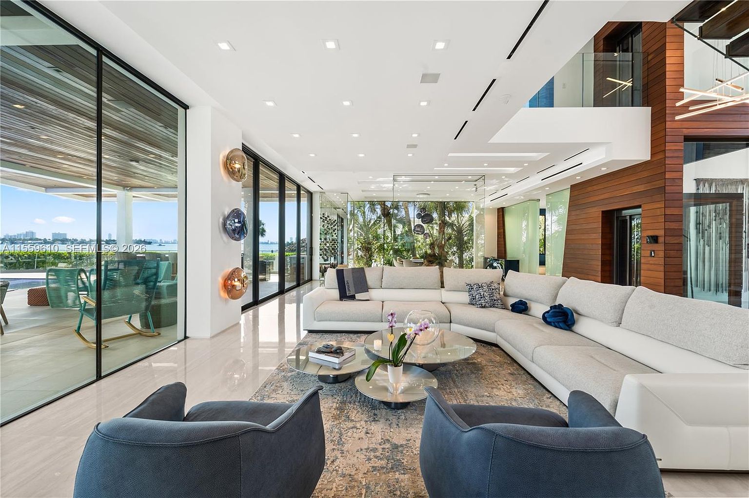 This is a spacious and luxurious living room featuring a large, sectional sofa, modern armchairs, and floor-to-ceiling windows offering stunning views. The room is decorated in a contemporary style with neutral colors and sleek finishes, creating an open and airy atmosphere. The polished floors and recessed lighting add to the elegance of the space.