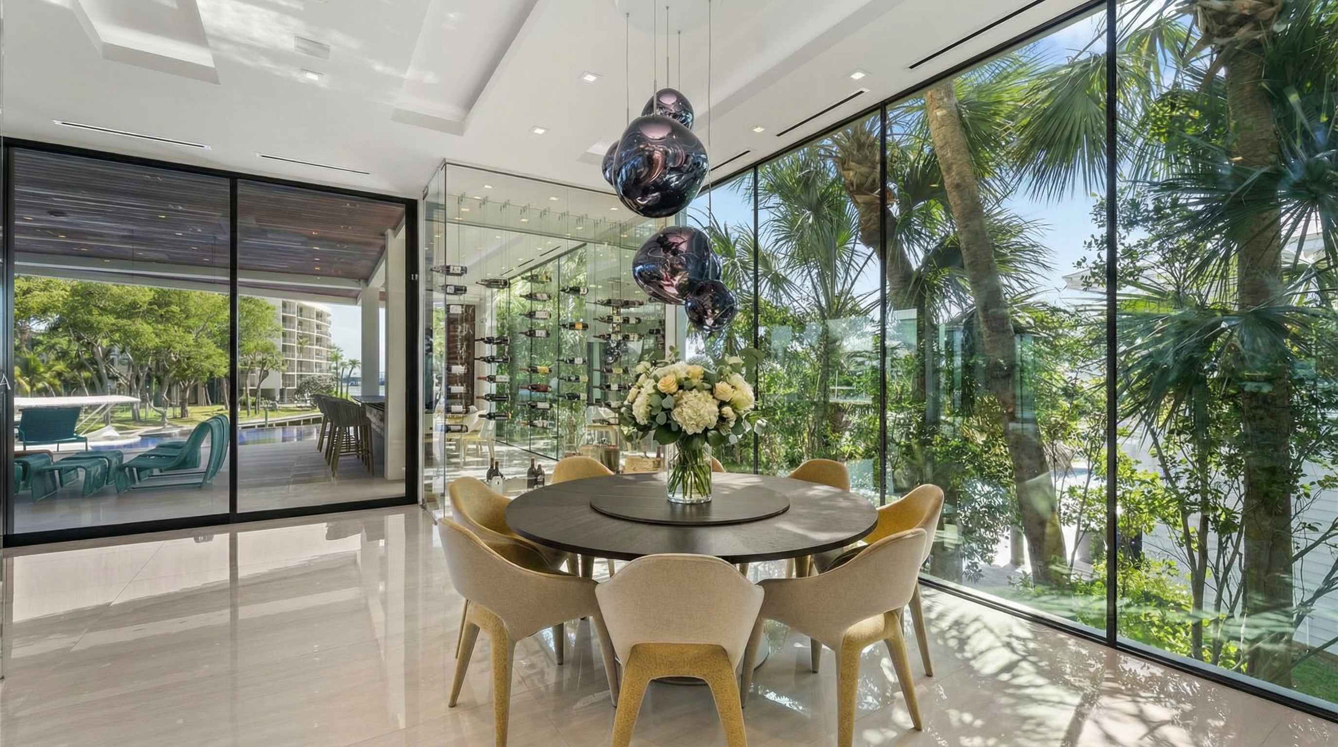 This modern dining area features a round, dark-finished table surrounded by stylish, contemporary chairs with textured upholstery. A stunning cluster of orb-shaped pendant lights hangs above a central floral arrangement, while floor-to-ceiling glass walls reveal an adjacent climate-controlled wine cellar and a lush outdoor view, emphasizing a bright and sophisticated atmosphere.