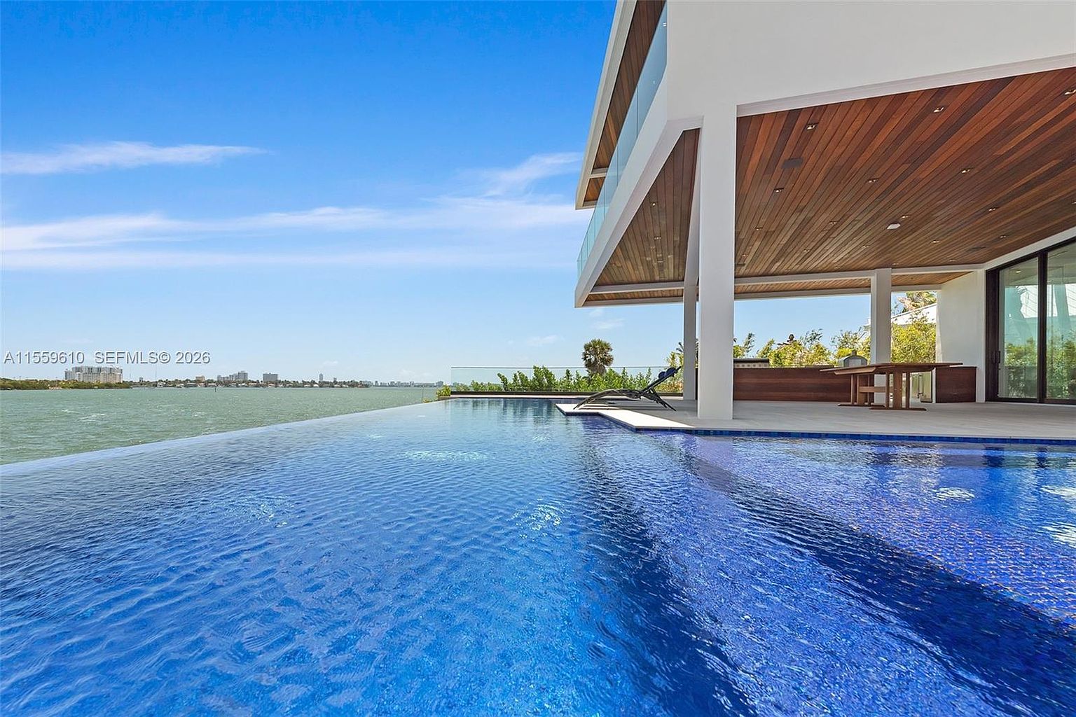 This image showcases a luxurious infinity pool that seamlessly blends with the ocean view. The modern architecture of the house features clean lines, a wooden ceiling, and glass railings, creating a sophisticated and inviting outdoor space. A lounge chair sits poolside, inviting relaxation and enjoyment of the stunning scenery.