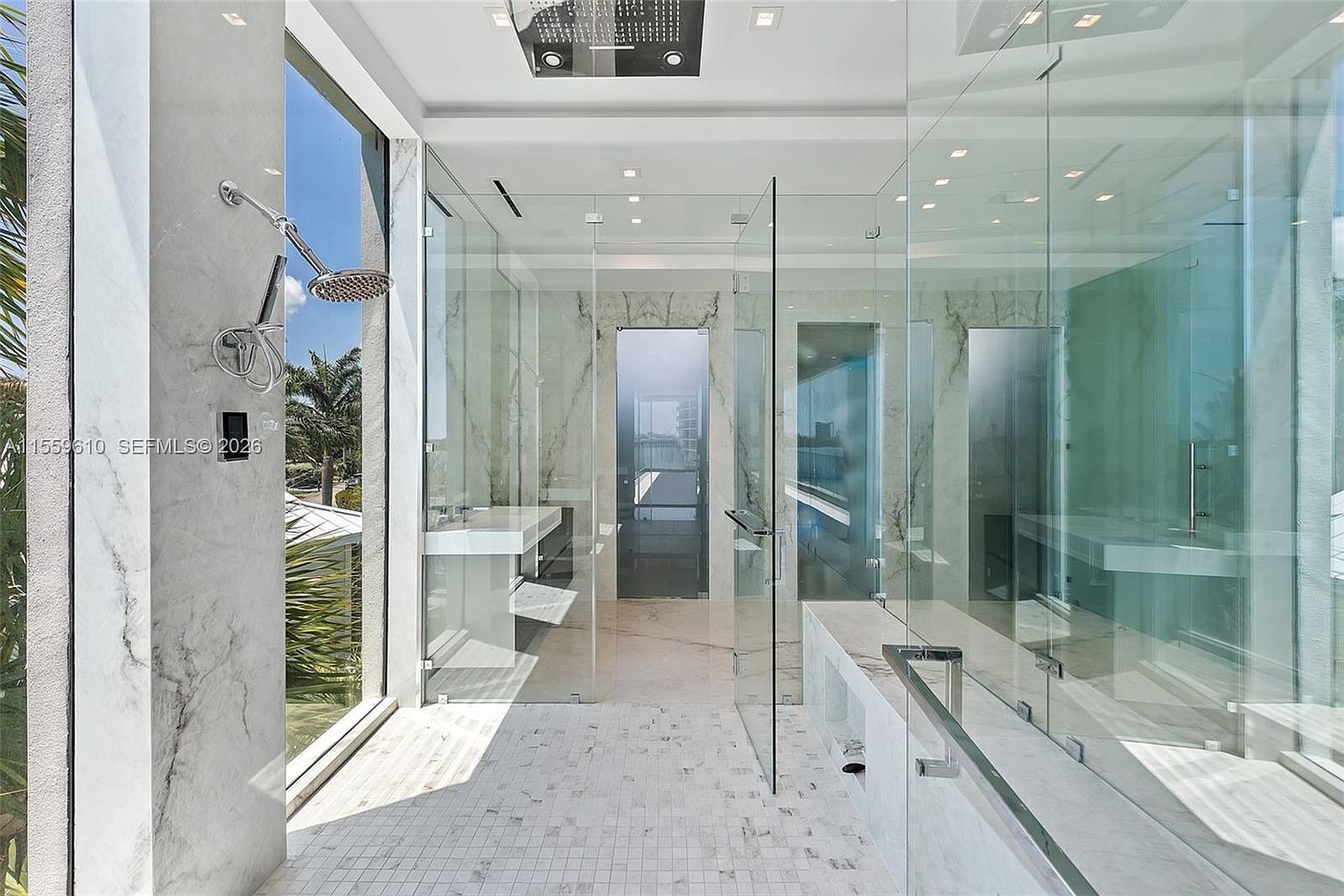 This is a luxurious primary bathroom featuring a spacious walk-in shower enclosed with clear glass panels, showcasing modern design and high-end finishes. The bathroom includes marble walls, a sleek vanity with a white countertop, and mosaic tile flooring, creating a bright and elegant atmosphere. The shower has a rainfall showerhead and a handheld showerhead.