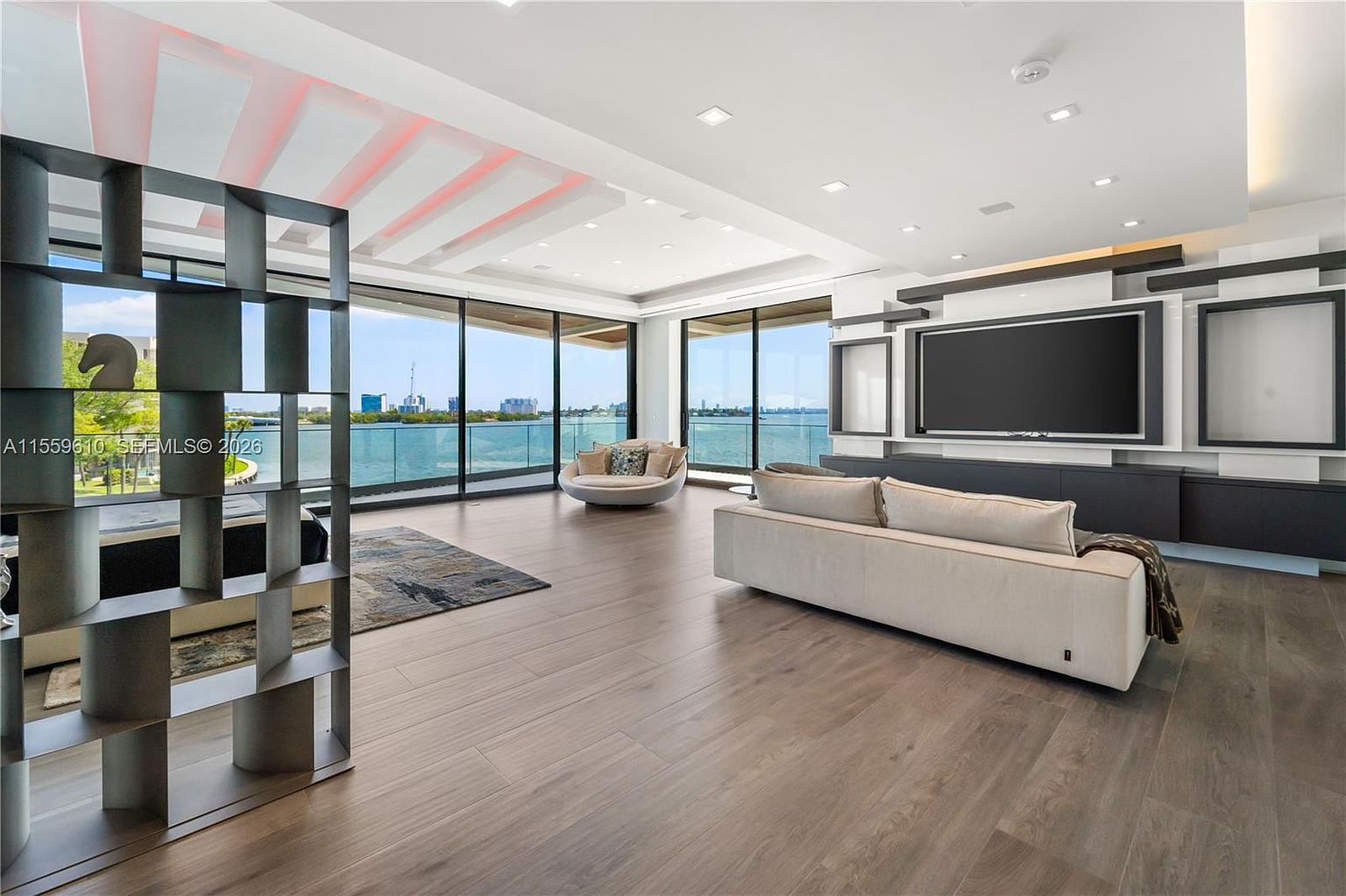 This is a spacious and modern living room with floor-to-ceiling windows offering a stunning waterfront view. The room features hardwood floors, a large television mounted on a custom wall unit, and contemporary furniture including a long sofa and a round chair. A decorative room divider adds visual interest and a touch of elegance.