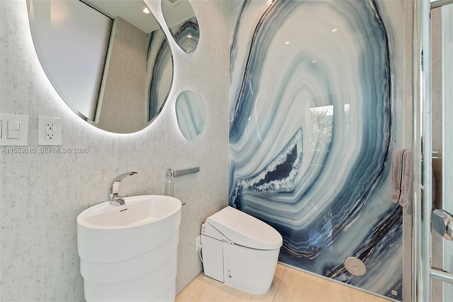 This is a modern bathroom featuring a unique pedestal sink with a chrome faucet, a sleek toilet, and a striking blue and white agate-inspired wall panel. The bathroom also includes a round, backlit mirror and smaller decorative mirrors, adding to the contemporary aesthetic. The overall impression is clean, stylish, and luxurious.