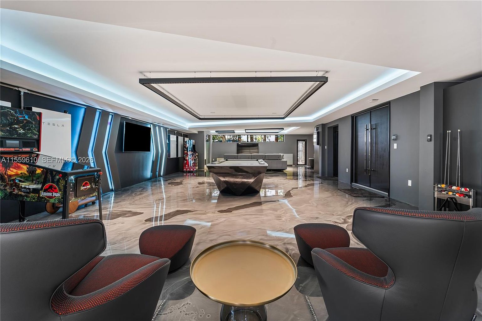 This is a spacious and modern living room featuring a unique pool table, arcade games, and a seating area with stylish chairs and a round table. The room is well-lit with recessed lighting and accent lighting, and the marble flooring adds a touch of luxury. The overall impression is one of entertainment and sophistication.