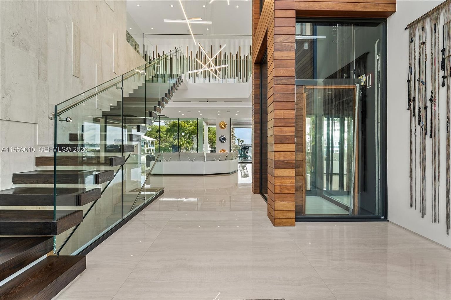 This interior shot showcases a grand foyer with a modern staircase featuring dark wood steps and glass railings. The open-concept design leads to a living area with a large white sectional sofa visible in the background. The space is characterized by clean lines, light-colored tile flooring, and a combination of natural wood and contemporary lighting fixtures, creating a luxurious and airy atmosphere.