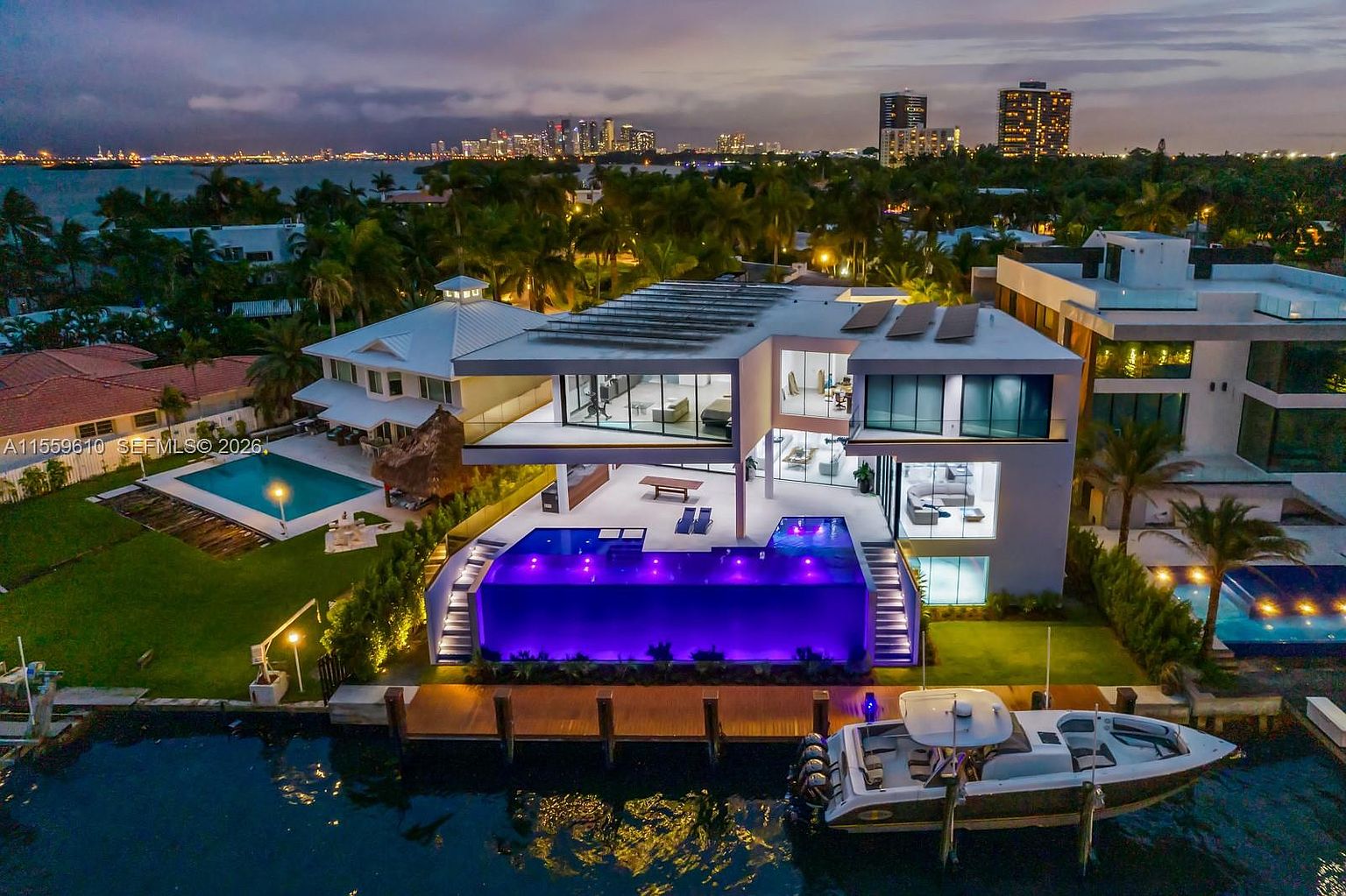This aerial view showcases a luxurious modern home with a striking architectural design. The property features a stunning infinity pool illuminated with purple lights, a private dock with a boat, and expansive outdoor living spaces. The backdrop includes a cityscape at dusk, adding to the property's allure and exclusivity.