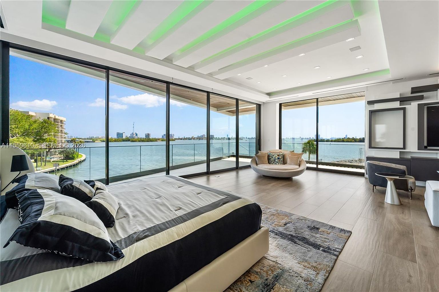 This is a luxurious primary bedroom featuring floor-to-ceiling windows with stunning waterfront views. The room is decorated in a modern style with a neutral color palette, accented by a black and white bedspread and a stylish area rug. The room also includes a seating area with a unique round sofa and a modern entertainment center.