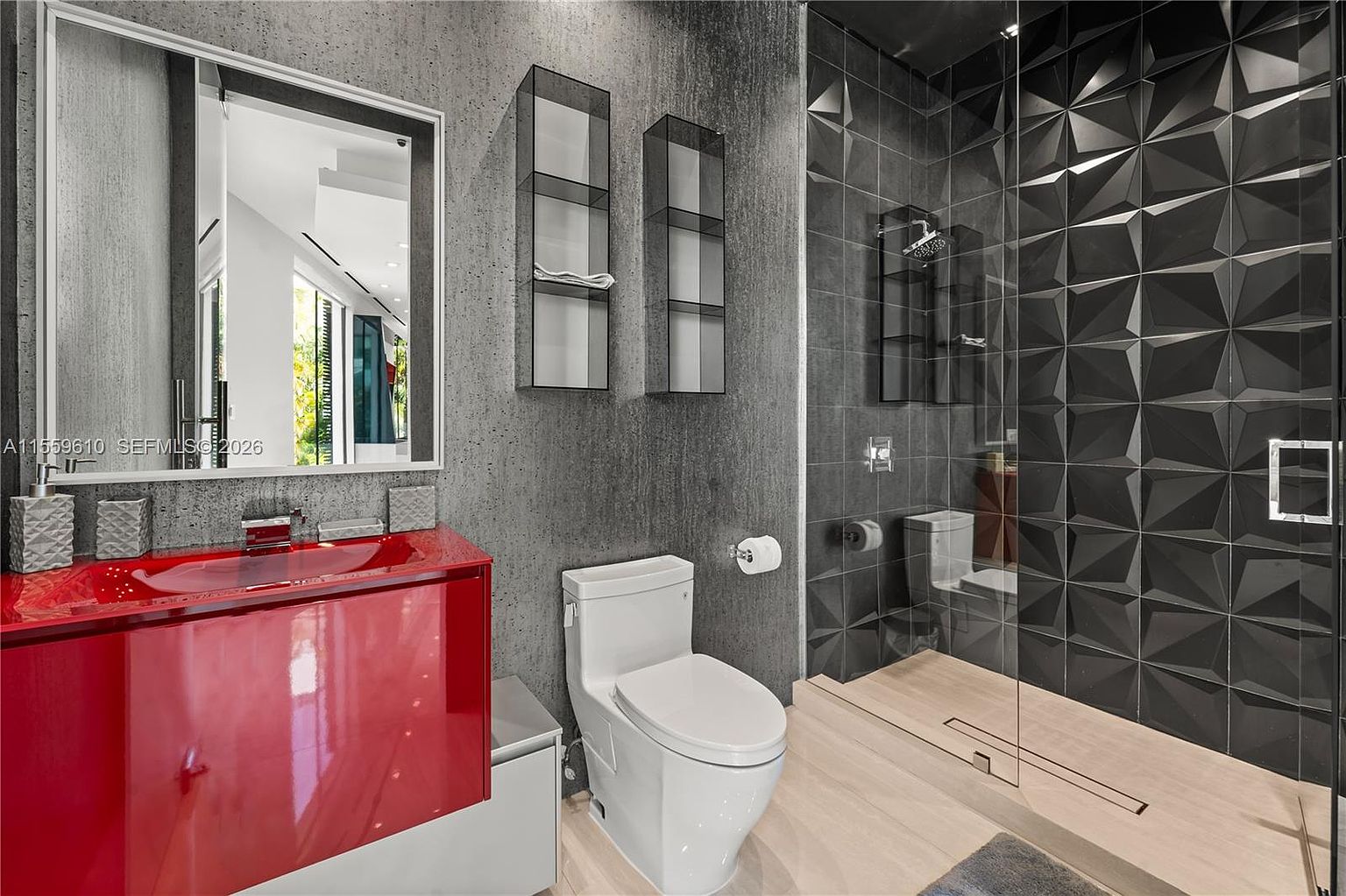 This is a modern primary bathroom featuring a red vanity with a unique design, a toilet, and a walk-in shower with textured black tiles. The walls are a textured gray, and the flooring appears to be light wood. The shower has a glass partition, and there are floating shelves on the wall next to the toilet.