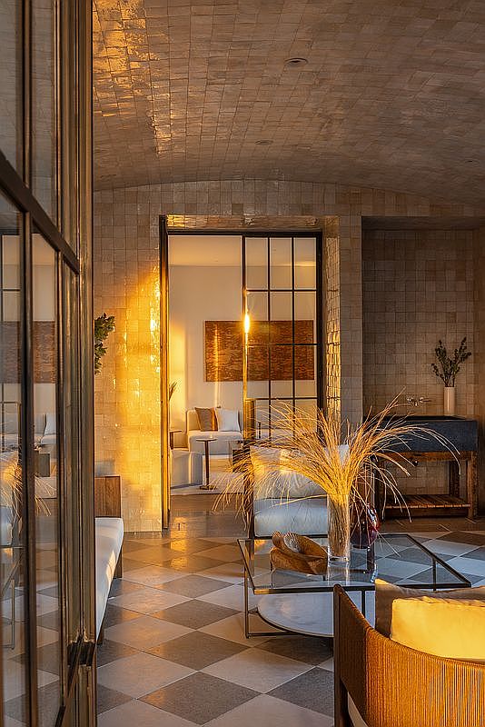 This sophisticated interior space features a barrel-vaulted ceiling covered in textured tiles and a striking checkerboard floor pattern. A glass-paned metal door provides a view into an adjacent room, while a modern glass coffee table and a decorative vase with tall dried grasses anchor the foreground. The warm, golden lighting creates an inviting and luxurious atmosphere, highlighting the rich textures of the stone walls and the elegant architectural design.