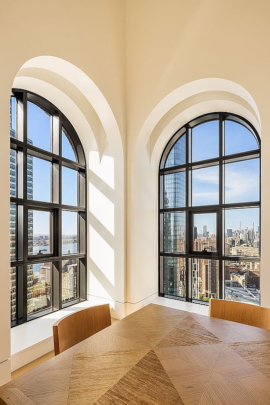 This bright dining area features a geometric patterned light wood table positioned in a corner with two large, arched windows. The windows offer expansive city views and are framed by clean, white walls that emphasize the height of the space. The minimalist design and natural light create an airy, sophisticated atmosphere suitable for a high-end urban residence.