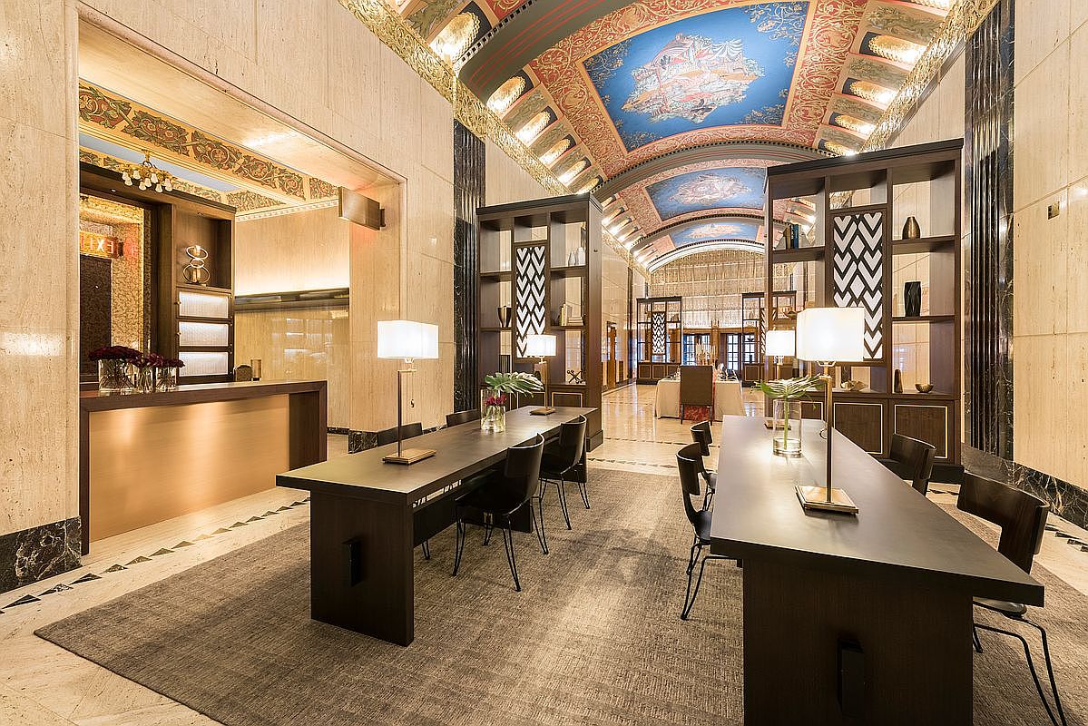 This grand, opulent lobby or lounge area features a high, arched ceiling adorned with intricate, colorful murals and decorative molding. The space is furnished with long, dark-toned tables paired with modern chairs, creating a sophisticated workspace or communal area. The room is accented by tall, open-shelving units with geometric patterns and warm, ambient lighting, conveying a sense of luxury and historic elegance.
