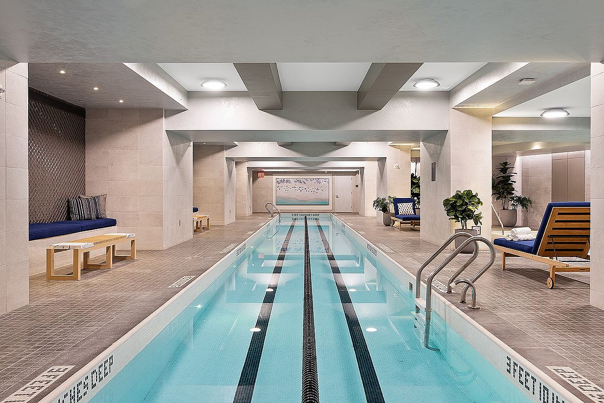 This indoor lap pool area features a long, narrow swimming lane with dark tile markers and a clean, modern aesthetic. The space is surrounded by neutral-toned tiled walls, recessed ceiling lighting, and comfortable lounge seating, including a cushioned bench and wooden lounge chairs. The perspective is centered down the length of the pool, emphasizing the symmetry and professional design of this high-end residential amenity.