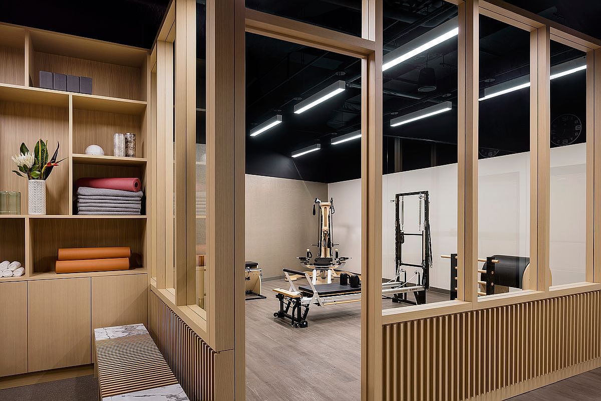 This professional-grade home gym features a dedicated workout space enclosed by light wood-framed glass partitions, offering a clean and modern aesthetic. The interior is equipped with specialized Pilates equipment, while the foreground includes a built-in shelving unit stocked with yoga mats, blocks, and a decorative vase. The perspective looks through the glass wall into the studio, highlighting the contrast between the warm wood tones and the dark, industrial-style ceiling.