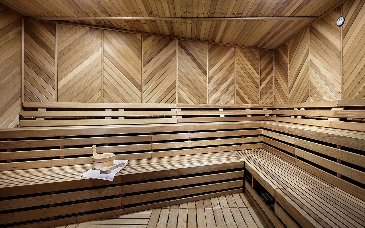 This interior view of a sauna features light wood paneling arranged in a sophisticated chevron pattern on the walls and ceiling. The space includes multi-level bench seating crafted from horizontal wood slats, with a wooden bucket and ladle resting on a white towel on the lower bench. A small circular thermometer is mounted on the upper wall, contributing to the clean, minimalist, and relaxing atmosphere of this wellness amenity.