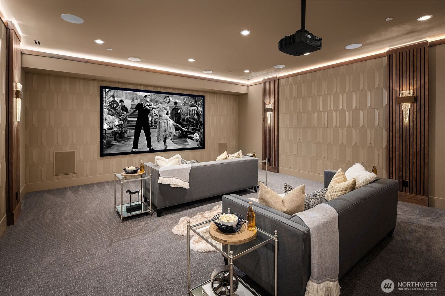 This is an interior shot of a home theater room. The room features two gray sofas facing a large screen, with side tables holding snacks and drinks. The walls are decorated with textured panels and wooden accents, and a projector is mounted on the ceiling, creating a cozy and immersive entertainment space.