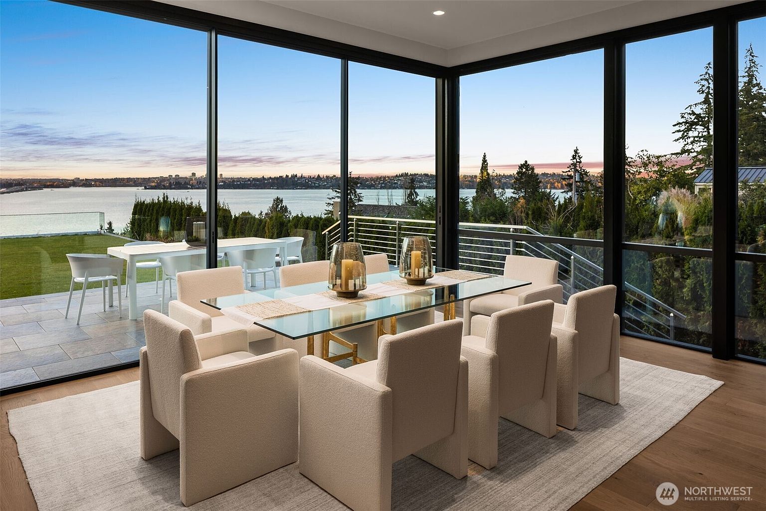 This is an interior shot of a dining room featuring a glass-top dining table with seating for ten. The room has floor-to-ceiling windows offering a panoramic view of a lake and landscape. The overall impression is one of modern elegance and spaciousness, ideal for entertaining and enjoying the scenic surroundings.