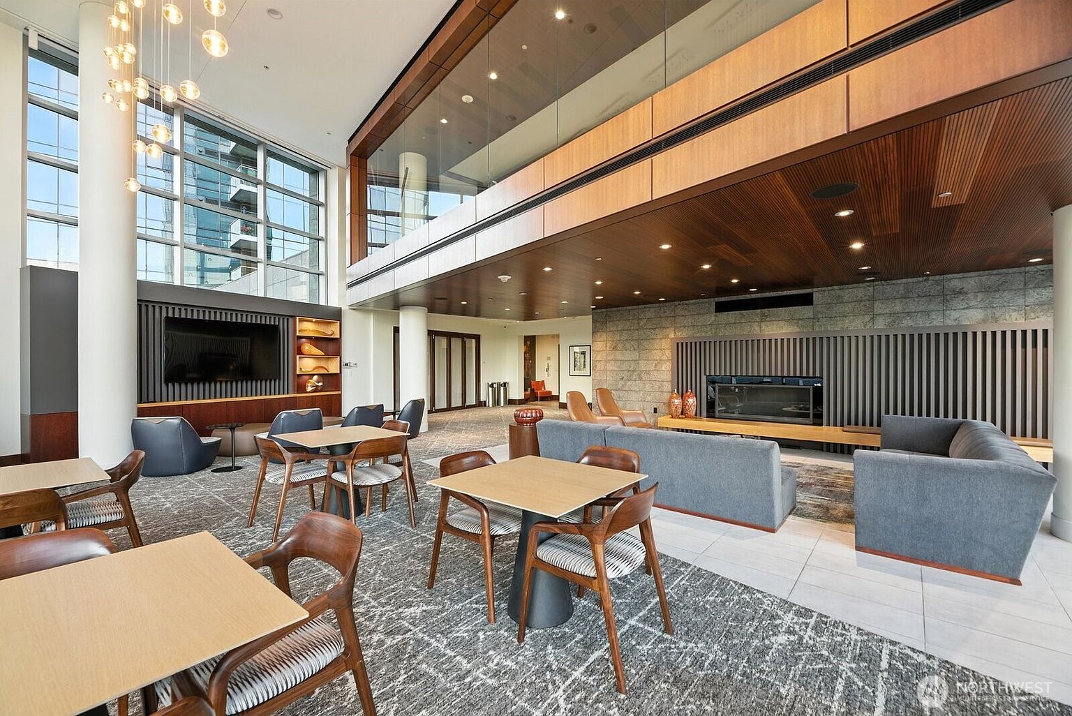 This is an interior shot of a modern clubhouse or amenity space. The area features a high ceiling, large windows, and a mix of seating arrangements including tables and chairs, as well as a gray sectional sofa near a fireplace. The design incorporates wood and stone elements, creating a sophisticated and inviting atmosphere.