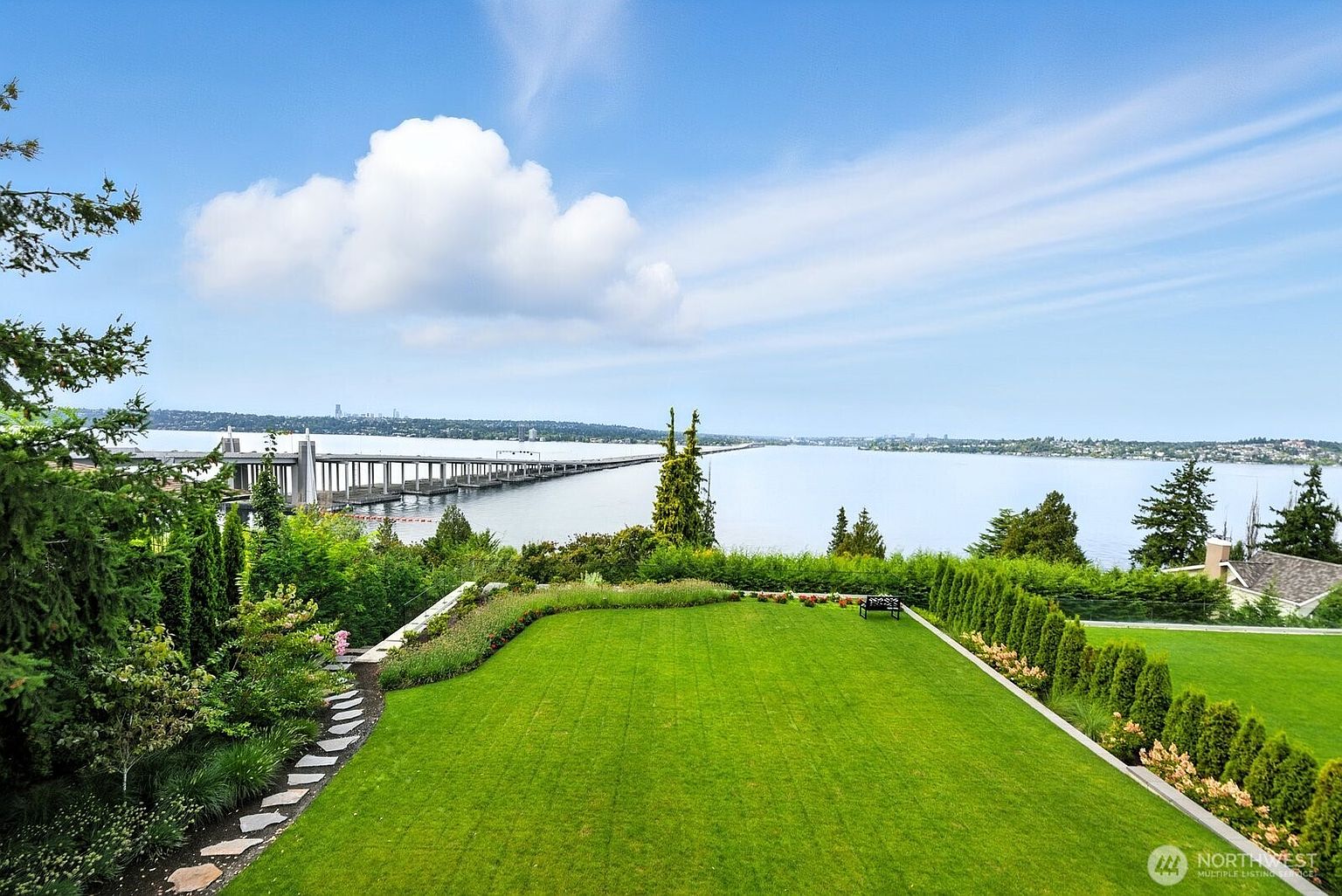 This image showcases a meticulously maintained lawn and garden with a stunning waterfront view. The lush green lawn is bordered by manicured hedges and flower beds, leading to a scenic vista of a bridge and the water. The perspective is from an elevated position, providing a comprehensive view of the property's outdoor space and its attractive landscaping.