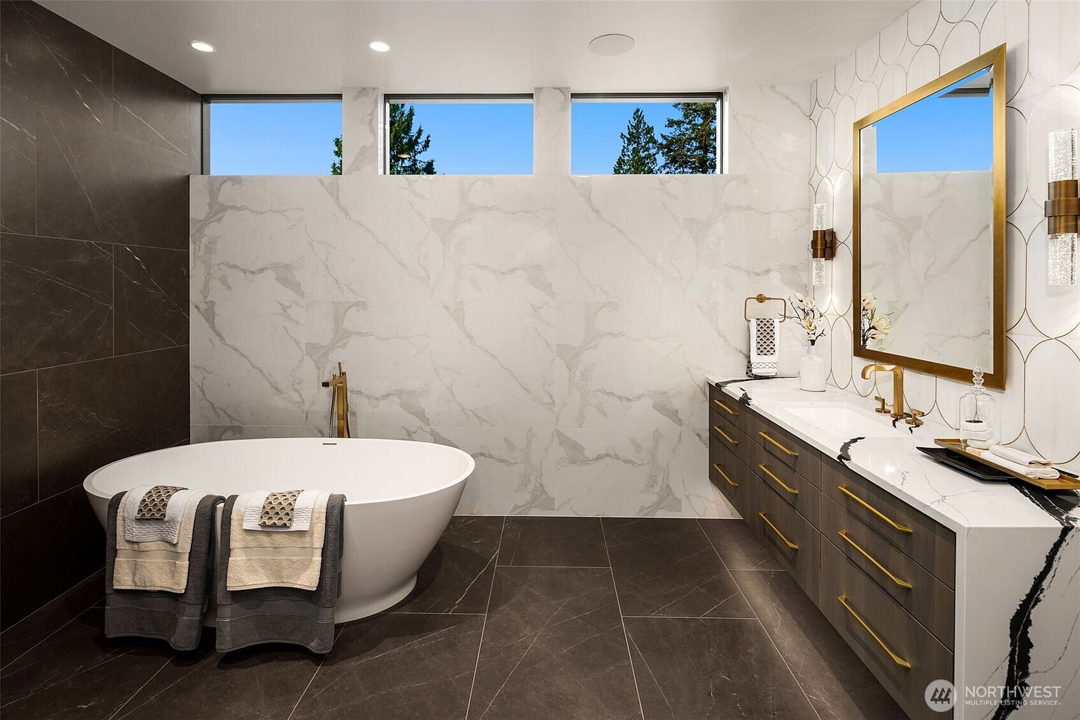 This is a luxurious primary bathroom featuring a freestanding soaking tub, a double vanity with modern fixtures, and elegant tile work. The color palette is neutral with gold accents, creating a spa-like atmosphere. Natural light streams in through the windows, enhancing the sense of tranquility and sophistication.
