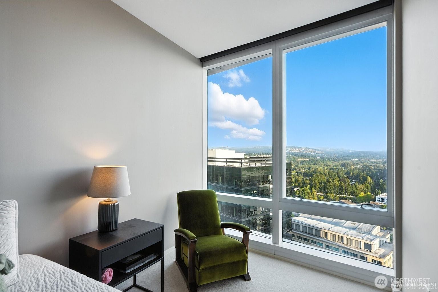 The image showcases a well-lit bedroom interior with a modern aesthetic. A green armchair sits near a large window offering a scenic view of the city and distant landscape. A bedside table with a lamp adds a cozy touch, while the overall impression is clean, bright, and inviting.