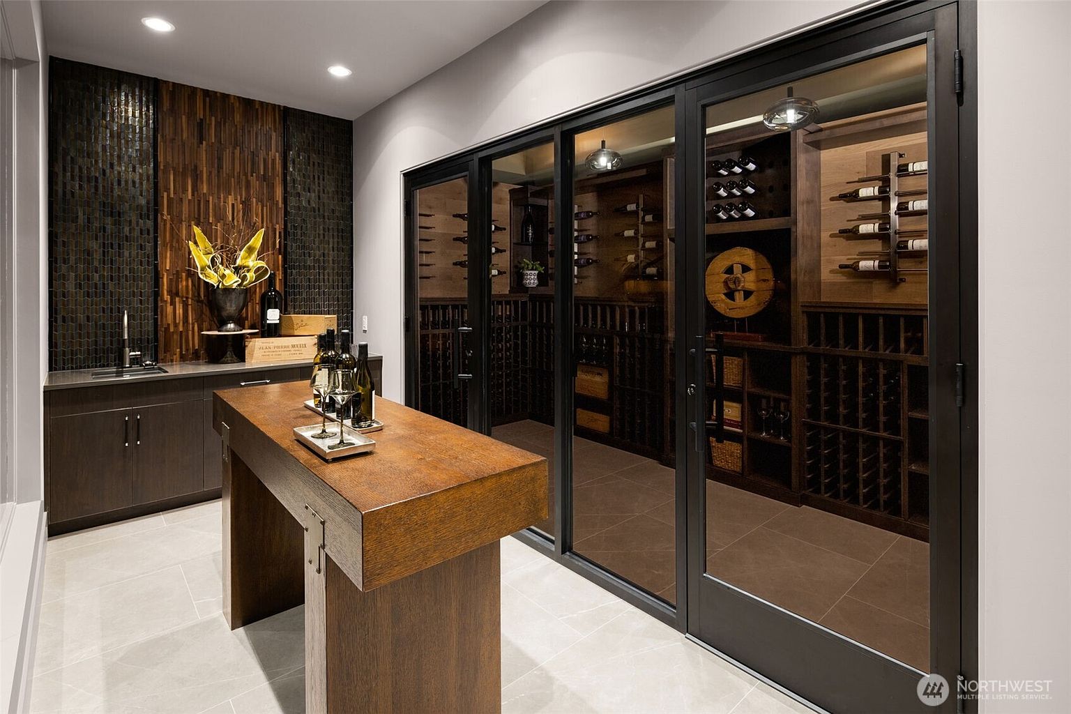 This image showcases a sophisticated wine cellar. The room features a custom-built wine rack system, a tasting table, and a bar area with a sink. The cellar is enclosed by glass doors, offering a view of the wine collection, and the overall design exudes luxury and elegance.