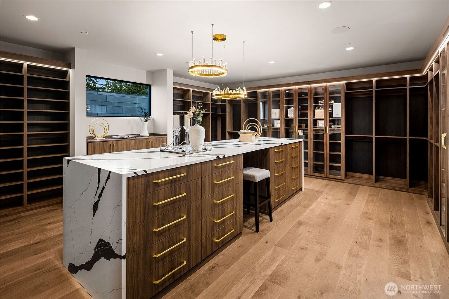 This is a luxurious walk-in closet featuring custom wood cabinetry with gold hardware and glass-fronted display cases. A large island with a marble countertop provides ample space for organization and display, while elegant chandeliers illuminate the space. The overall impression is one of high-end design and functionality.