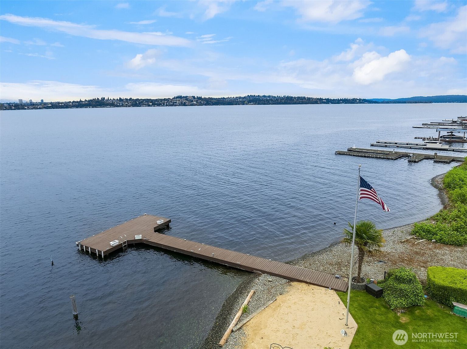 This aerial view showcases a waterfront property with a private dock extending into the lake. The property features a sandy beach area, a well-maintained lawn, and mature landscaping. The American flag is prominently displayed, adding a patriotic touch to the serene lakeside setting.