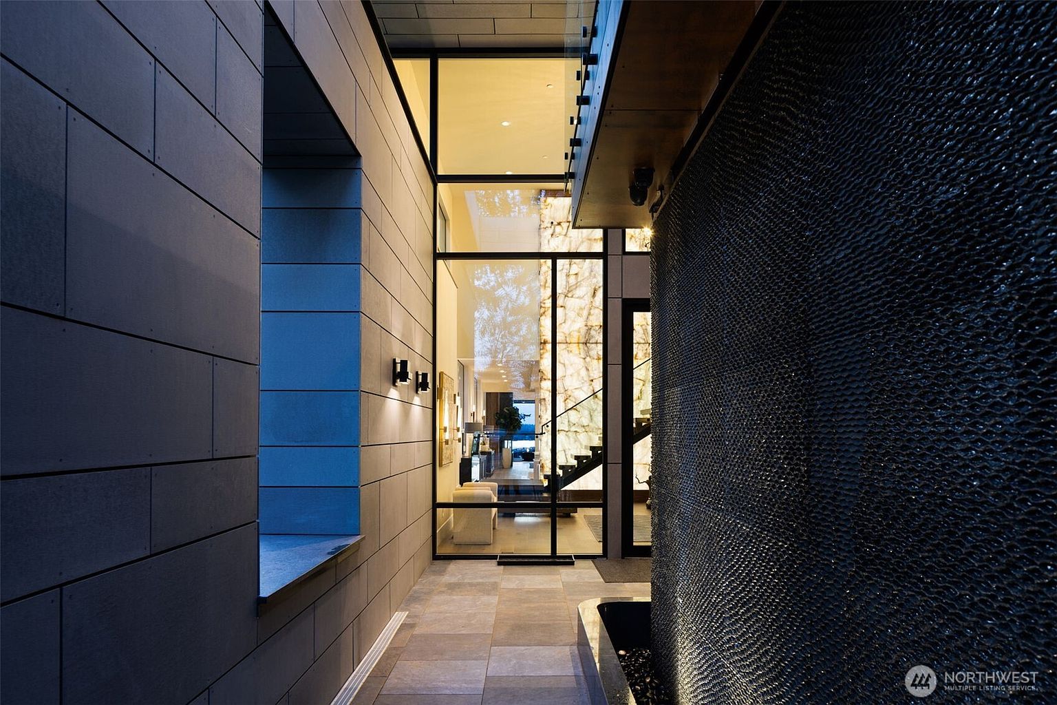 This image showcases a modern entryway with a striking water feature on the right and a textured wall on the left. The entrance features large glass panels and a door, providing a glimpse into the interior with stairs and a seating area. The flooring is tiled, and the overall design exudes contemporary elegance and sophistication.