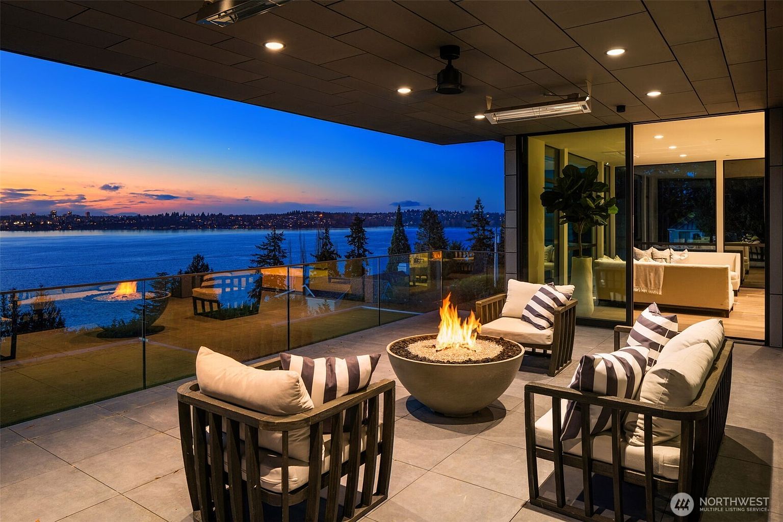 This image showcases a luxurious outdoor patio or deck area with a stunning waterfront view at dusk. The space features modern outdoor furniture arranged around a fire pit, creating a cozy and inviting atmosphere. The patio is enclosed by a glass railing, providing unobstructed views of the water and the city skyline in the distance, while the interior space is visible through large glass doors, seamlessly blending indoor and outdoor living.