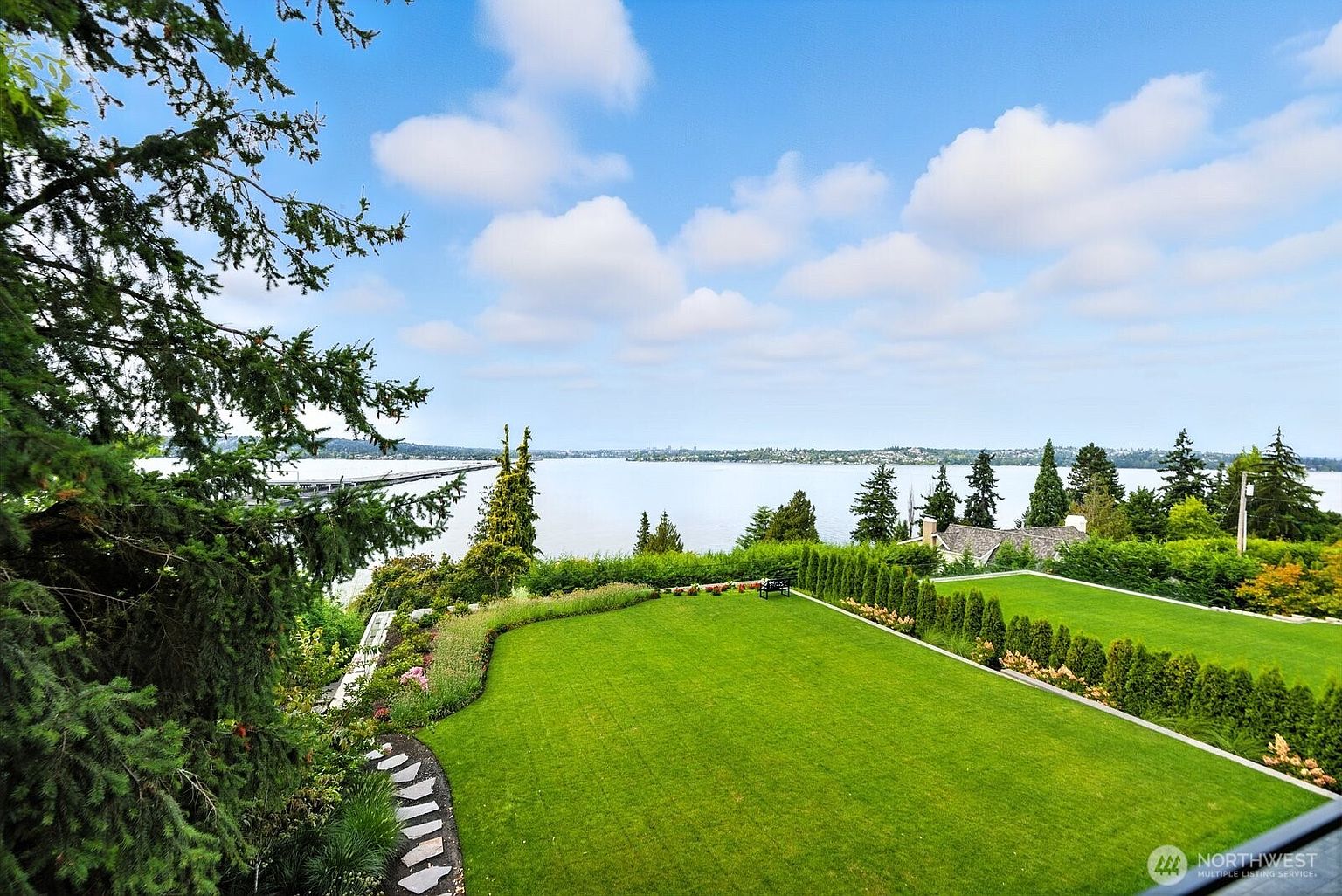This image showcases a meticulously maintained yard and garden with a stunning view of the water. The lush green lawn is bordered by manicured hedges and flower beds, creating a serene and inviting outdoor space. A stone pathway adds a touch of elegance, leading through the garden area.