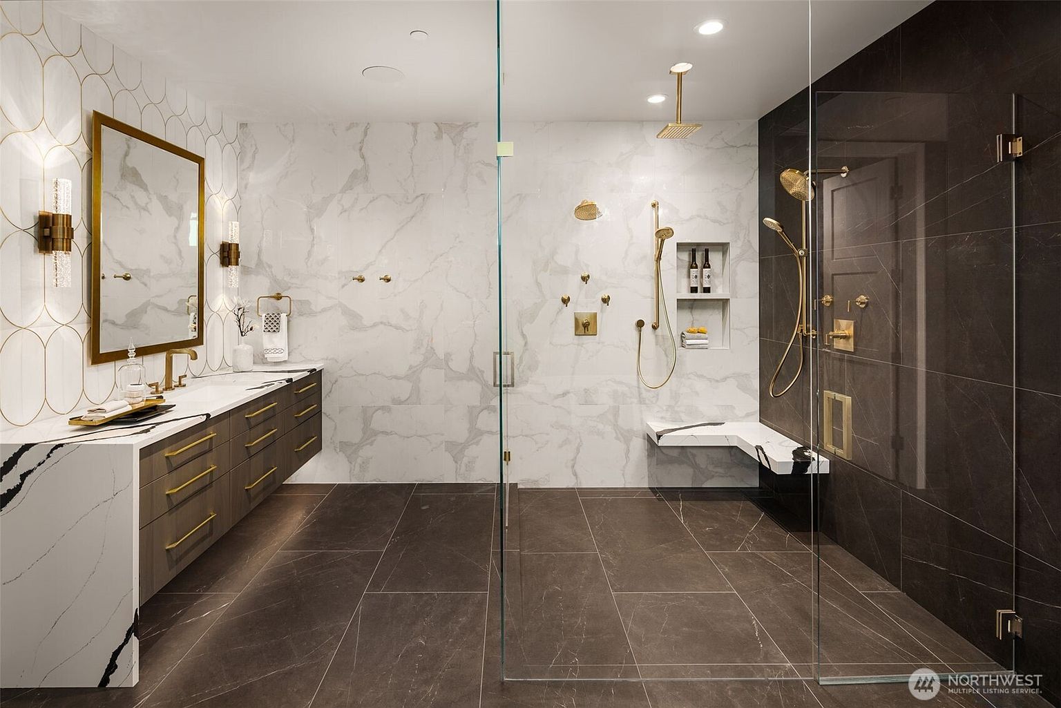 This is a luxurious primary bathroom featuring a double vanity with a marble countertop and gold fixtures. A glass-enclosed shower with both rainfall and handheld showerheads is visible, along with a built-in bench and niche for toiletries. The flooring is a dark tile, and the walls are a combination of white marble and dark, textured tiles, creating a modern and elegant space.