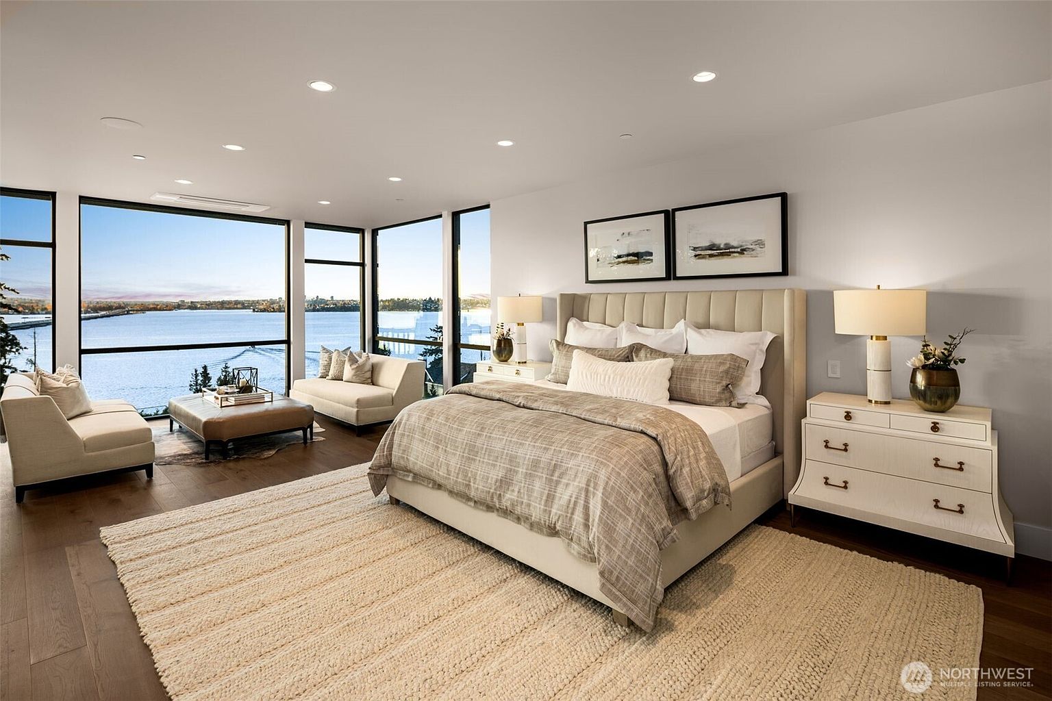 This is a bright and spacious primary bedroom featuring large windows with a waterfront view. The room is furnished with a king-size bed, a nightstand with a lamp, and a seating area with two chairs and a coffee table. The decor is modern and elegant, with neutral colors and natural light creating a serene atmosphere.