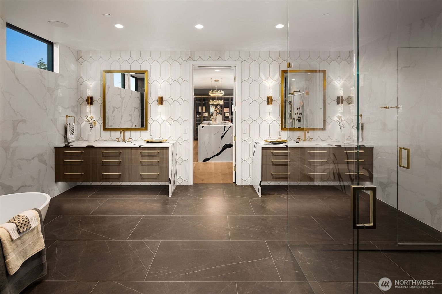 This is a luxurious primary bathroom featuring dual vanities with marble countertops and bronze fixtures. The walls are adorned with a combination of marble and patterned tiles, creating a sophisticated and elegant atmosphere. A glass-enclosed shower is visible on the right, and a glimpse into a walk-in closet can be seen through the doorway.