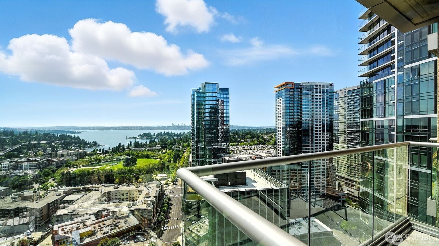 This image showcases a stunning view from a high-rise balcony, featuring modern glass railings and a glimpse of the city skyline. The perspective captures the surrounding cityscape, including other high-rise buildings, lush greenery, and a distant body of water under a partly cloudy sky. The overall impression is one of luxury and urban living.