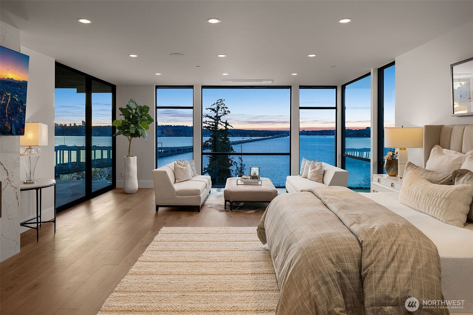 This is a luxurious primary bedroom featuring floor-to-ceiling windows with a stunning water view. The room is furnished with a large bed, two sofas, and a central ottoman, creating a comfortable and elegant living space. The hardwood floors and neutral color palette enhance the room's serene and upscale ambiance.