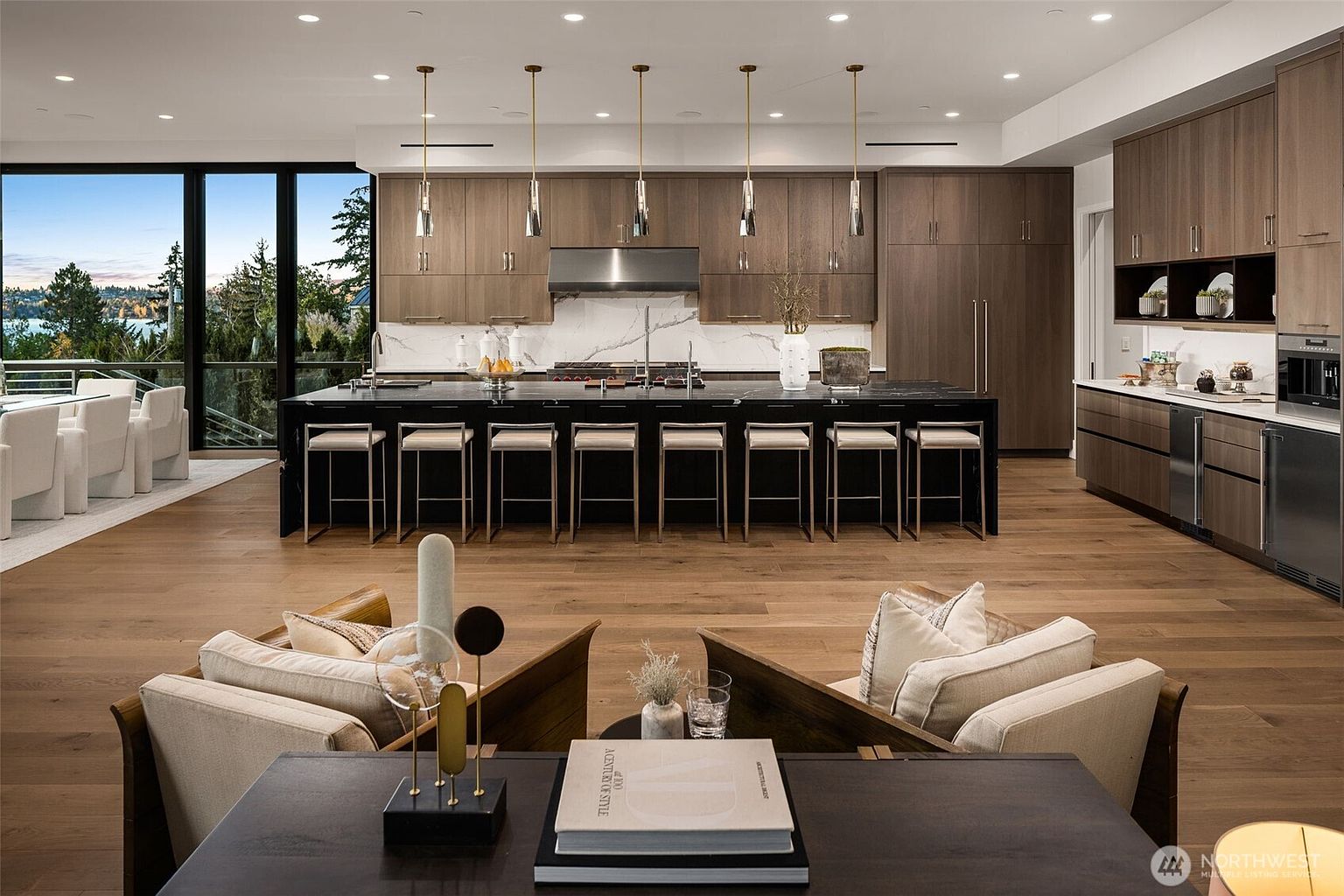 This is a wide-angle shot of a modern, open-concept kitchen and living area. The kitchen features a large black island with seating, wood cabinetry, stainless steel appliances, and pendant lighting. The living area includes comfortable seating and a view of the outdoors through large windows, creating a seamless indoor-outdoor living experience.