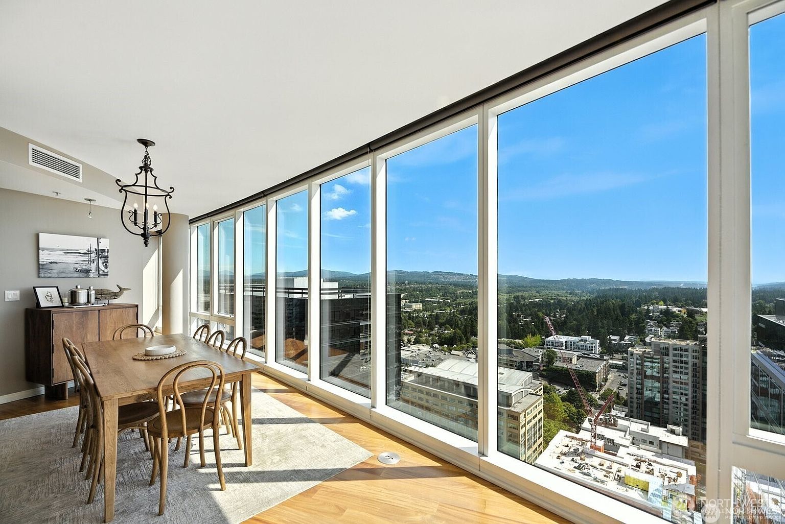 This is an interior shot of a dining room featuring a long wooden table with chairs, set on a patterned rug. A modern chandelier hangs above the table, and a wooden cabinet sits against the wall. Large windows offer a panoramic view of the city and surrounding landscape, creating a bright and airy atmosphere.