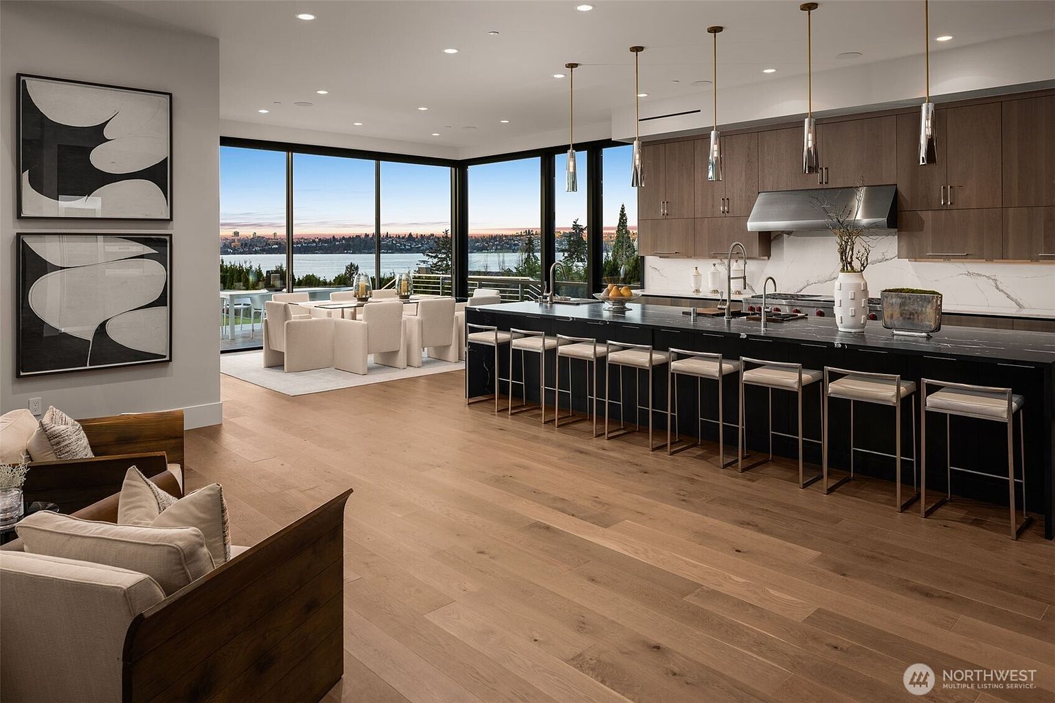 This is a spacious, modern kitchen with a large island featuring bar seating and dark countertops. The kitchen boasts sleek, dark wood cabinetry and stainless steel appliances. Large windows offer a stunning view of the water, and the open floor plan seamlessly connects the kitchen to the dining area and living space, creating an inviting atmosphere.